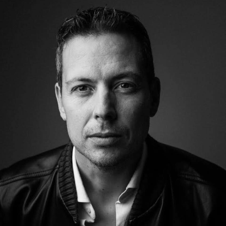 Black-and-white portrait of a man with short hair and stubble, wearing a leather jacket over a white collared shirt. Based in Siena, the wedding photographer in Italy is looking directly at the camera with a neutral expression against a plain, dark background. Tuscan Wedding Photographer