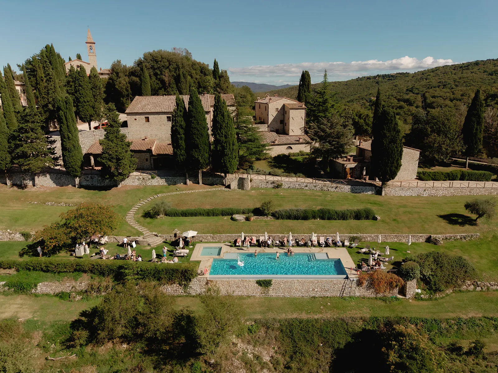 Aerial view of a countryside estate with stone buildings and tall cypress trees. In the foreground, there is a rectangular outdoor swimming pool surrounded by lounge chairs and people relaxing. Green hills and gardens fill the background under a clear sky. Tuscan Wedding Photographer