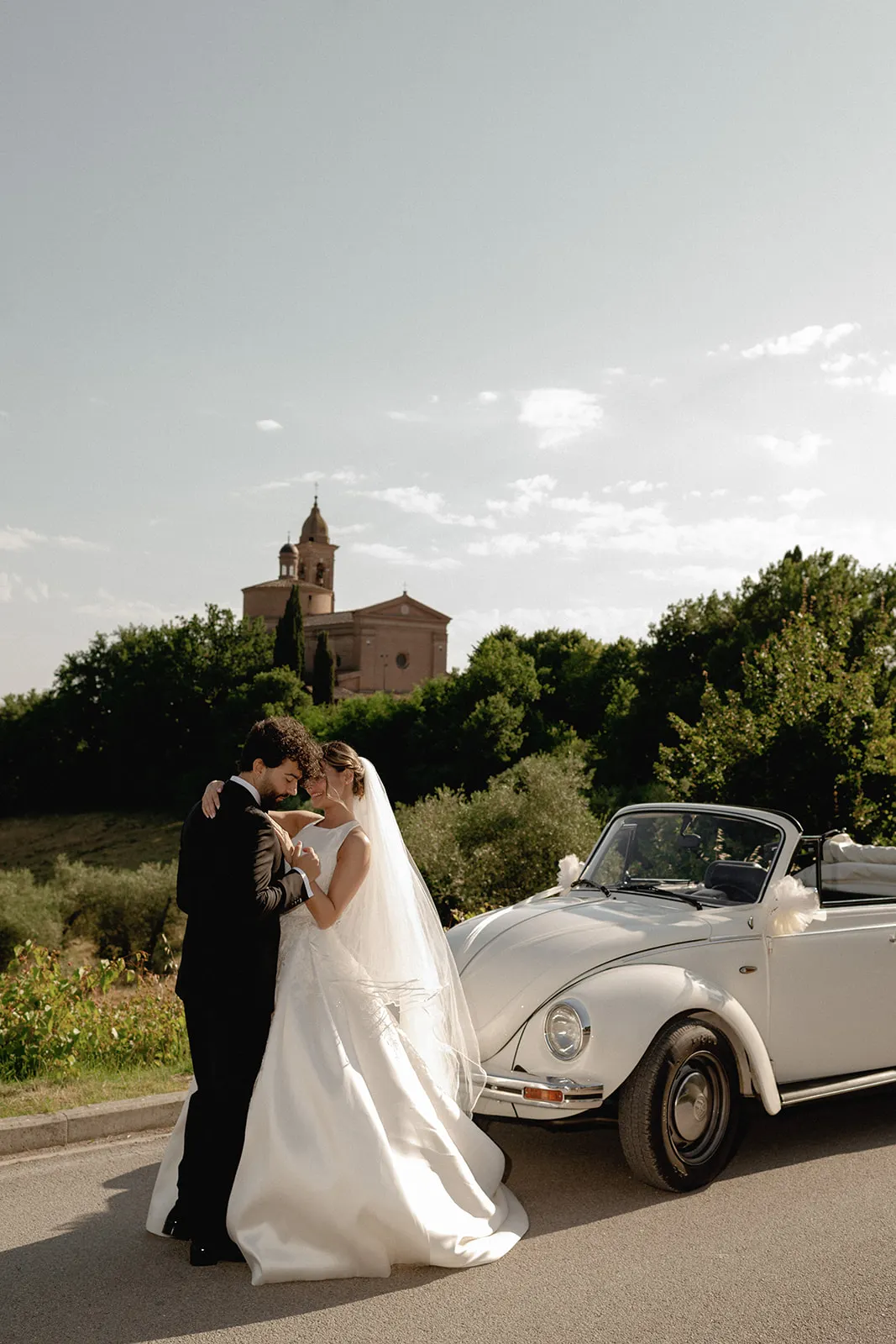 A bride and groom embrace beside a white vintage convertible on a sunlit road. Captured by a wedding photographer in Tuscany, the scene features trees and a brick church with a dome under a clear sky. Tuscan Wedding Photographer
