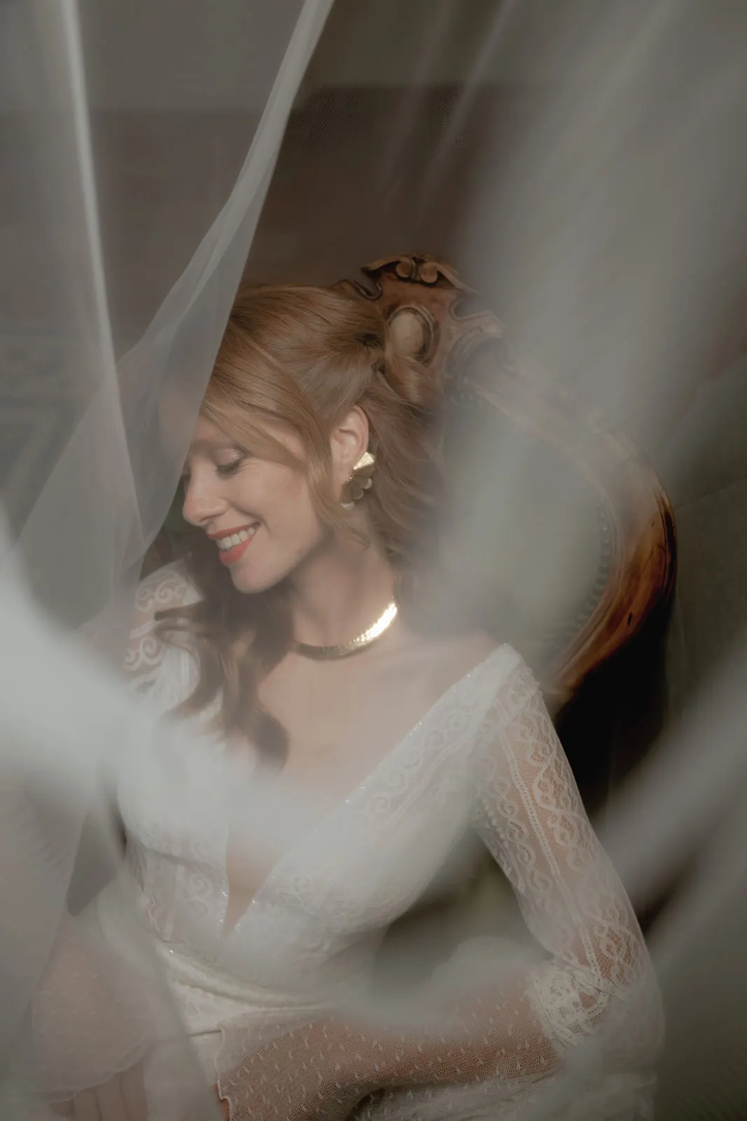 A smiling woman in a lace bridal gown sits on an ornate chair, eyes closed. Soft white veil fabric drapes around her, creating a dreamy, romantic effect. She wears gold earrings and a necklace, with wavy red hair styled back. Tuscan Wedding Photographer
