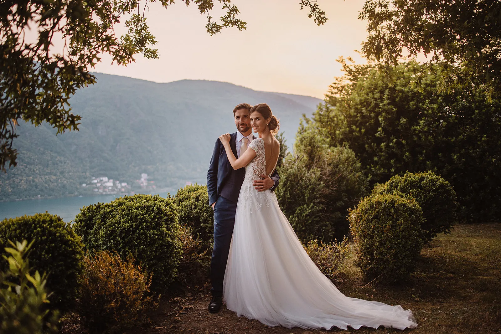 A bride in a flowing white gown and a groom in a dark suit stand embraced outdoors at sunset, surrounded by greenery. Captured by a wedding photographer Tuscany, mountains and water gleam softly behind them under the evening sky. Tuscan Wedding Photographer