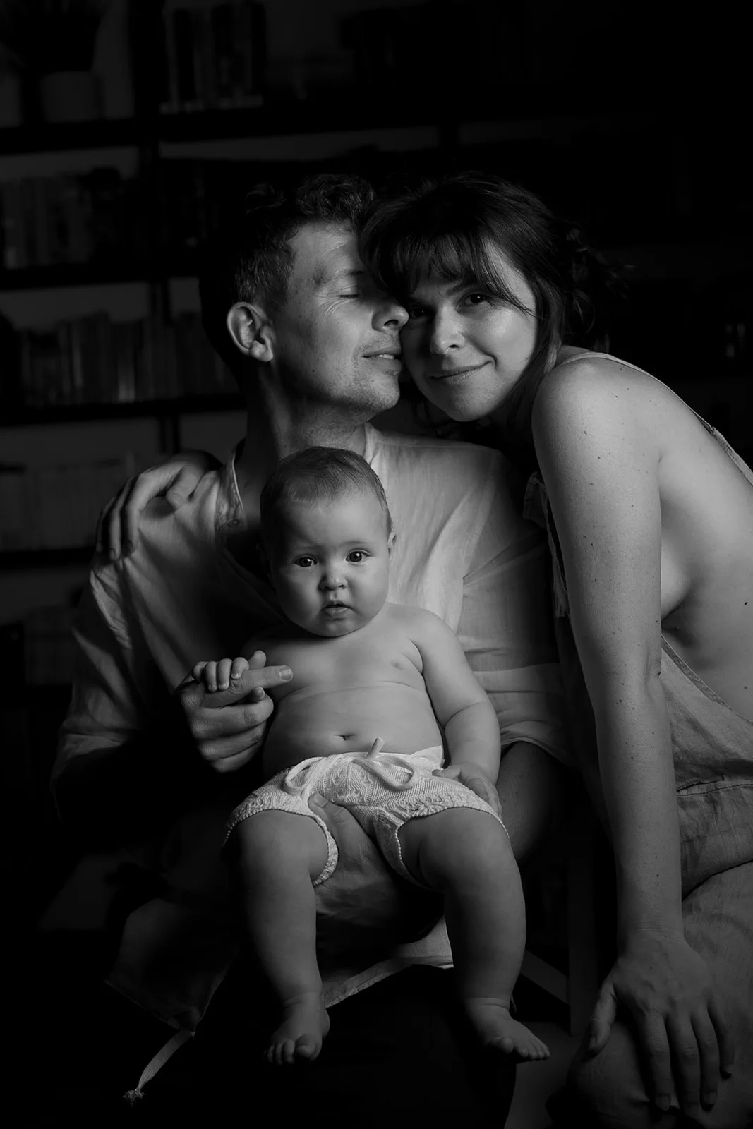 A black-and-white photo of a smiling couple holding a baby, softly lit with shelves in the background. Captured by Federico Pannacci, wedding photographer based in Siena, it beautifully reflects warmth and connection. Tuscan Wedding Photographer