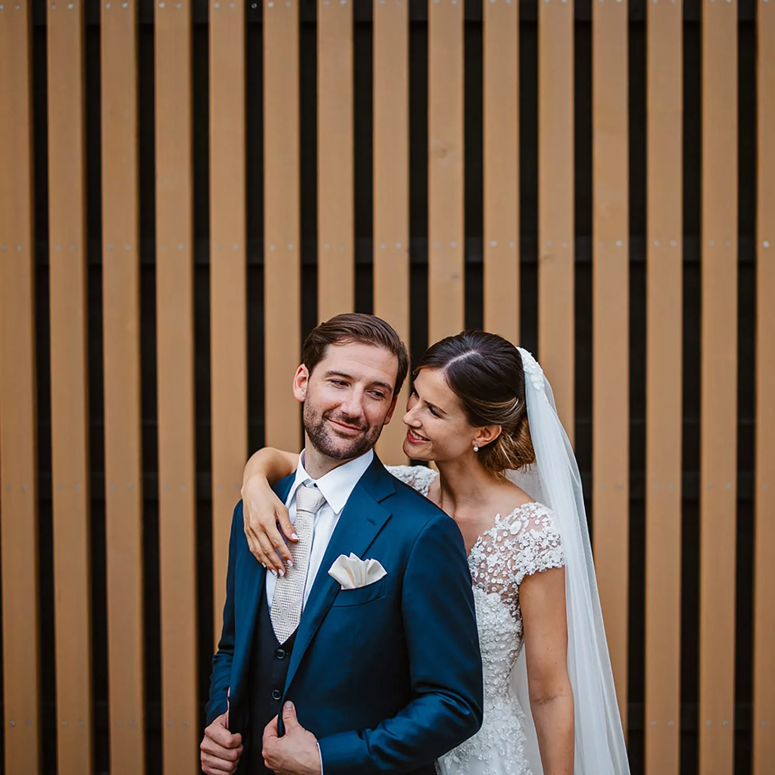 A bride in a lace wedding dress and veil smiles at her groom in a blue suit with a white tie and pocket square. Captured by a wedding photographer Tuscany, they stand close together before vertical wooden slats, her arm around his shoulders. Tuscan Wedding Photographer