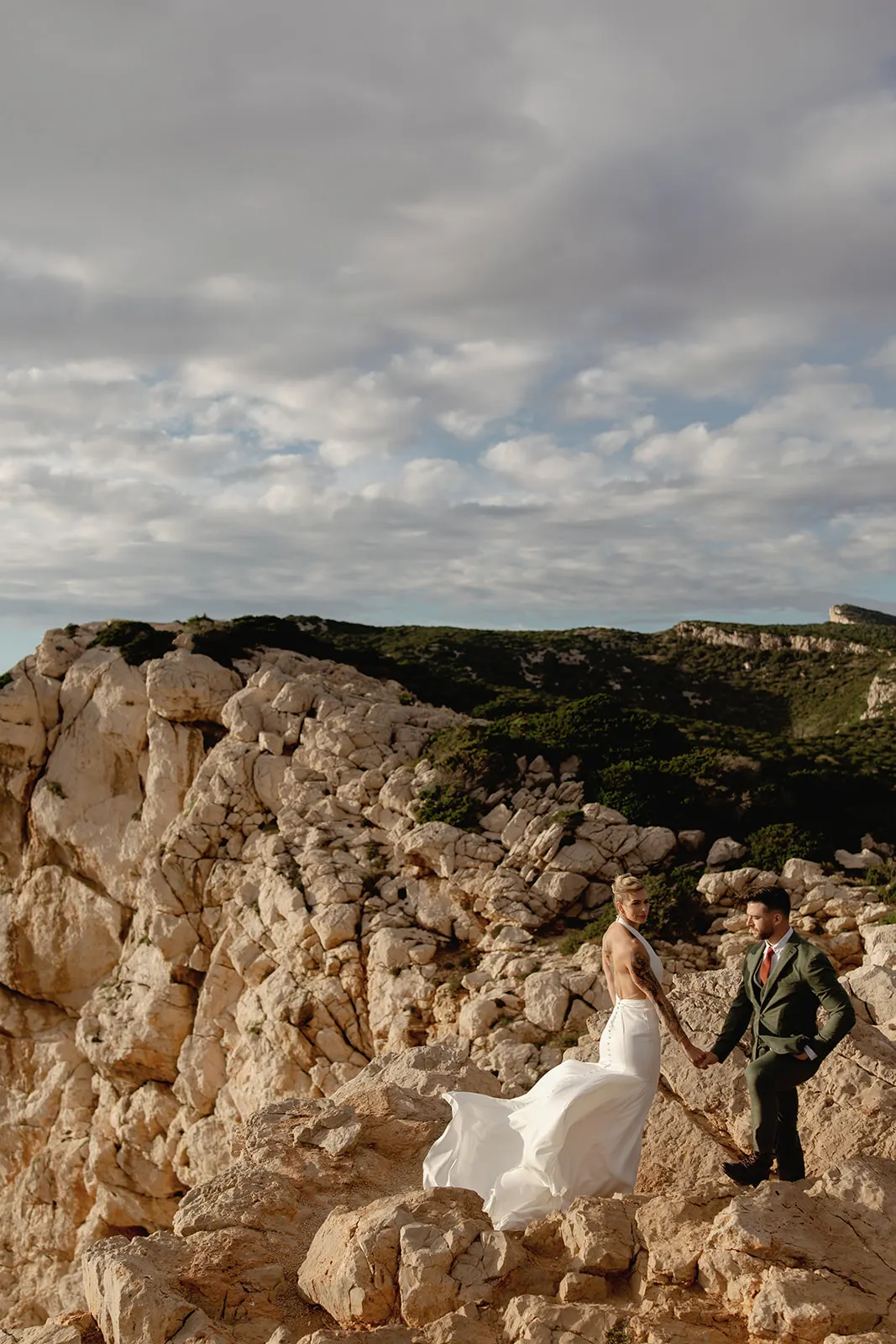 A couple in formal wedding attire stands on rocky terrain with dramatic beige cliffs and green hills, captured by a wedding photographer Tuscany. They hold hands beneath a partly cloudy sky, evoking a romantic, adventurous mood. Tuscan Wedding Photographer
