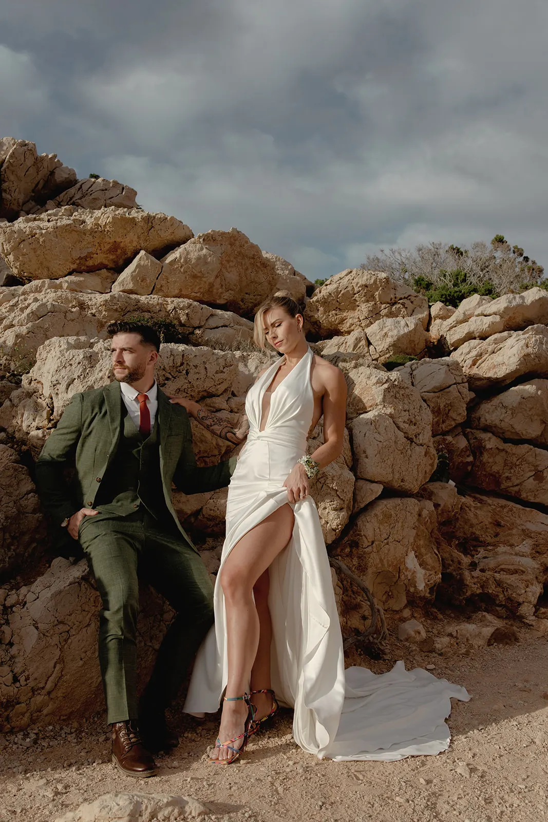 A couple poses by large rocky cliffs. The man sits in a green suit and red tie, gazing to the side. The woman stands confidently in a white halter-neck gown, hand on hip, both under a cloudy sky—captured by a wedding photographer Tuscany style. Tuscan Wedding Photographer