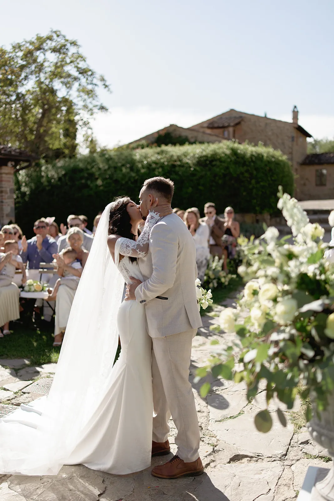 A bride and groom share a kiss outdoors during their ceremony, captured beautifully by a wedding photographer Tuscany. The bride’s white gown and veil, the groom’s light suit, and lush greenery create an unforgettable moment with guests watching amidst white flowers. Tuscan Wedding Photographer