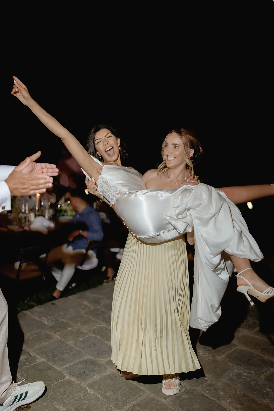 Two women smile joyfully at night; one in a white dress and heels is lifted by the other, who wears a light pleated skirt. The lifted woman raises an arm in celebration—an unforgettable moment captured by a wedding photographer in Tuscany. Tuscan Wedding Photographer