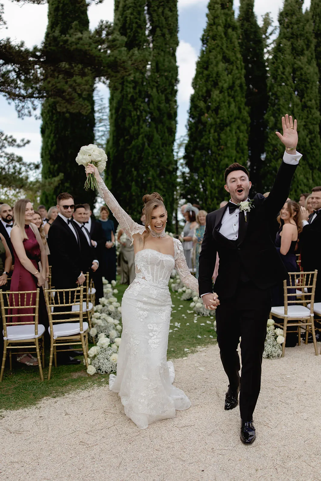 A joyful bride in a lace wedding dress holds a bouquet aloft, smiling beside a groom in a black tuxedo who waves as they walk down an outdoor aisle lined with guests, golden chairs, and tall green trees in the background. Tuscan Wedding Photographer