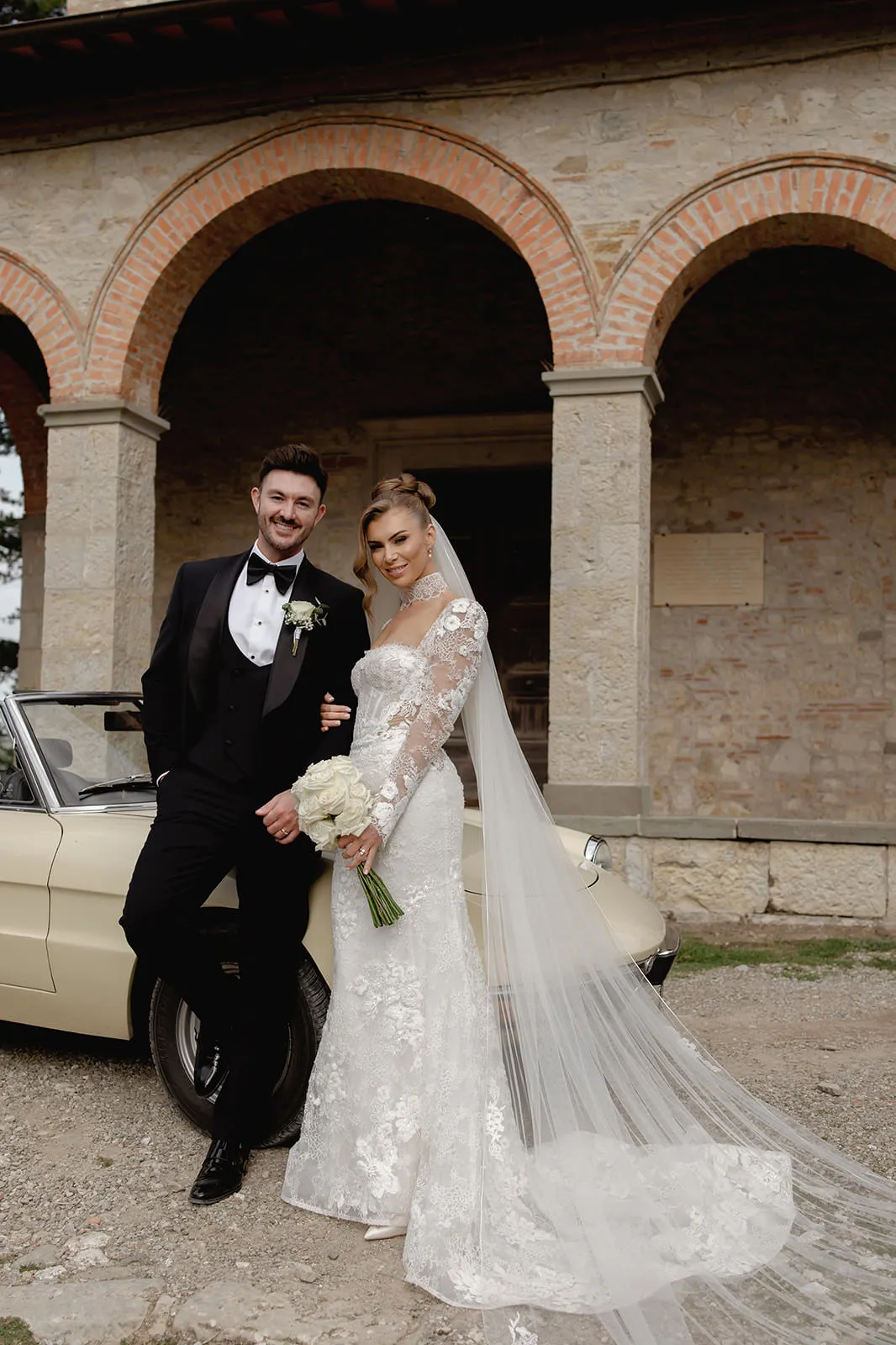 A bride in a white lace dress with a long veil and holding a bouquet of white flowers stands beside a groom in a black tuxedo. They smile in front of a vintage car and a stone building with arches and brick details. Tuscan Wedding Photographer