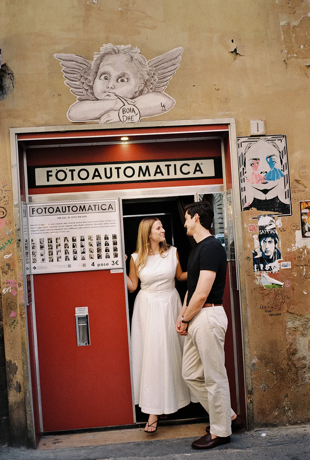 A woman in a white dress and a man in a black shirt stand smiling at each other in front of a red FOTOAUTOMATICA photo booth on a graffiti-marked wall—perfect inspiration for any wedding photographer Tuscany could offer. Tuscan Wedding Photographer