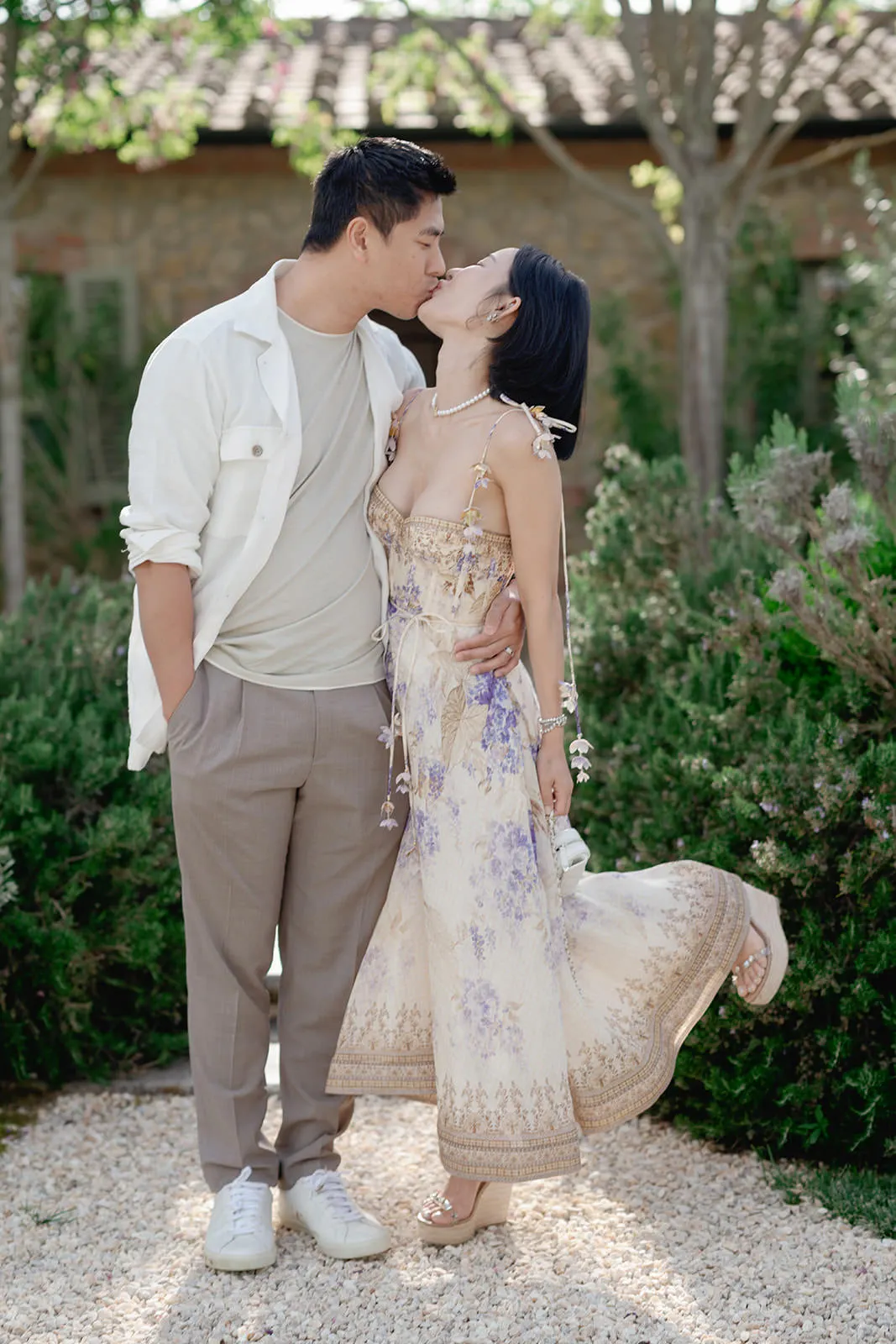 A couple stands outside, kissing. The man wears a white shirt over a beige t-shirt, taupe pants, and white sneakers. The woman, in a flowing floral sundress and heels, lifts one leg as she leans into him. They are surrounded by greenery and trees. Tuscan Wedding Photographer