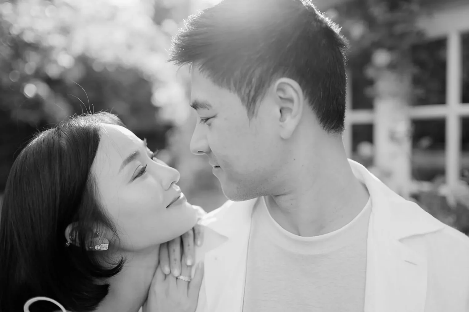 A black-and-white photo of a couple outdoors. They gaze into each others eyes and smile softly. The woman gently holds the man’s shoulder, showing an engagement ring on her finger. Sunlight creates a soft glow, and greenery is visible in the blurred background. Tuscan Wedding Photographer