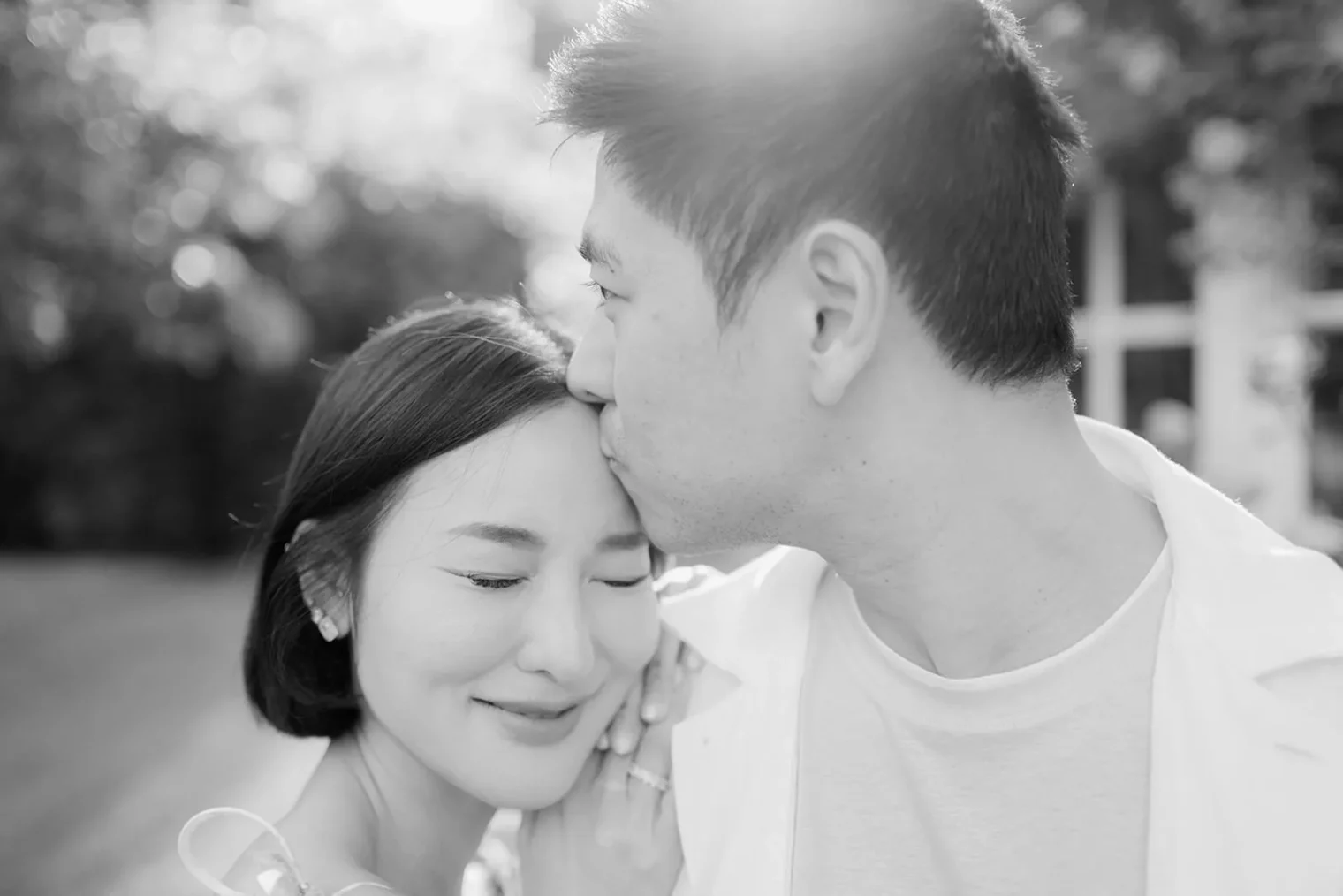 A man gently kisses a smiling womans forehead outdoors. She has her eyes closed and is holding his arm, both appearing peaceful and content. Sunlight softly brightens the background, creating a warm, intimate mood. The image is in black and white. Tuscan Wedding Photographer