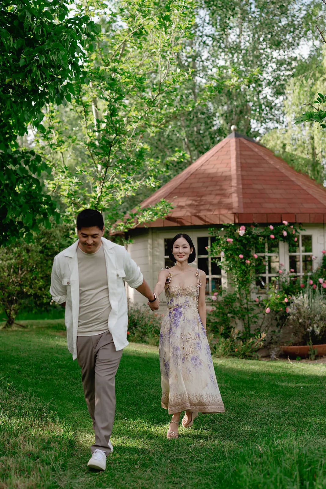 A man and woman hold hands, walking on green grass in a garden. The woman wears a floral dress; the man wears light pants and a white shirt. Behind them is a small, round house with a red roof, white windows, and flowers climbing the walls. Trees surround the scene. Tuscan Wedding Photographer