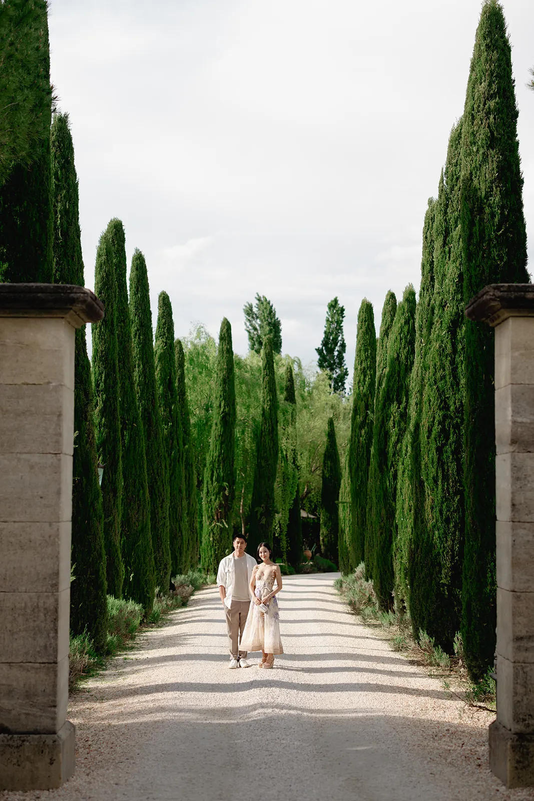 A couple walks down a wide gravel path lined with tall, lush cypress trees, framed by stone pillars on either side. The sky above is mostly cloudy, and the green trees create a dramatic, elegant corridor effect. Tuscan Wedding Photographer