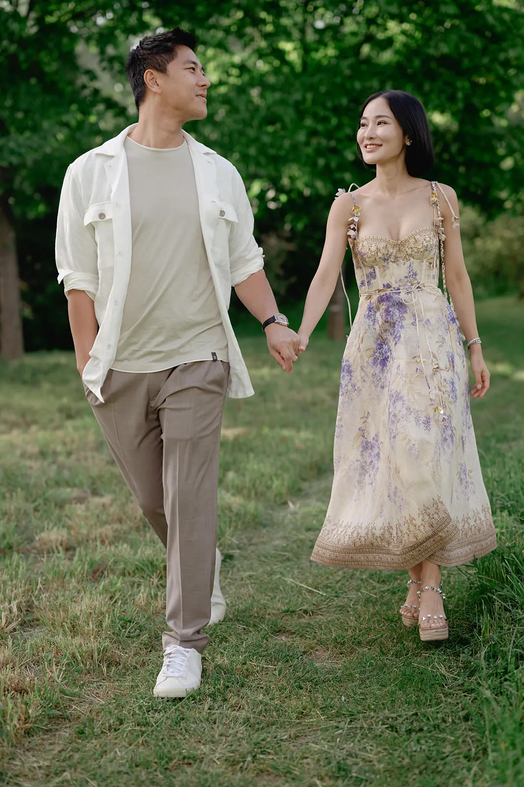 A young couple walks hand in hand on grass, both smiling at each other. The man wears a light shirt, beige pants, and white sneakers. The woman wears a light, floral-patterned dress and sandals. Green trees fill the background, indicating a park or garden setting. Tuscan Wedding Photographer