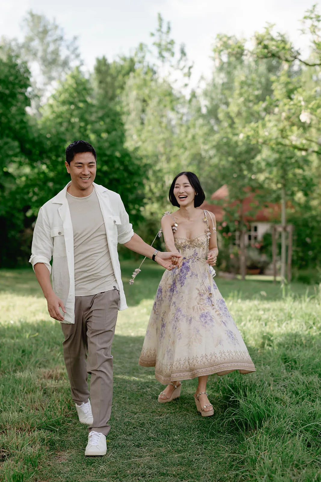 A smiling couple walks hand in hand on a grassy path outdoors. The man wears a light shirt, pants, and sneakers. The woman wears a light, floral, sleeveless dress and sandals. They appear happy, surrounded by green trees and sunlight. Tuscan Wedding Photographer