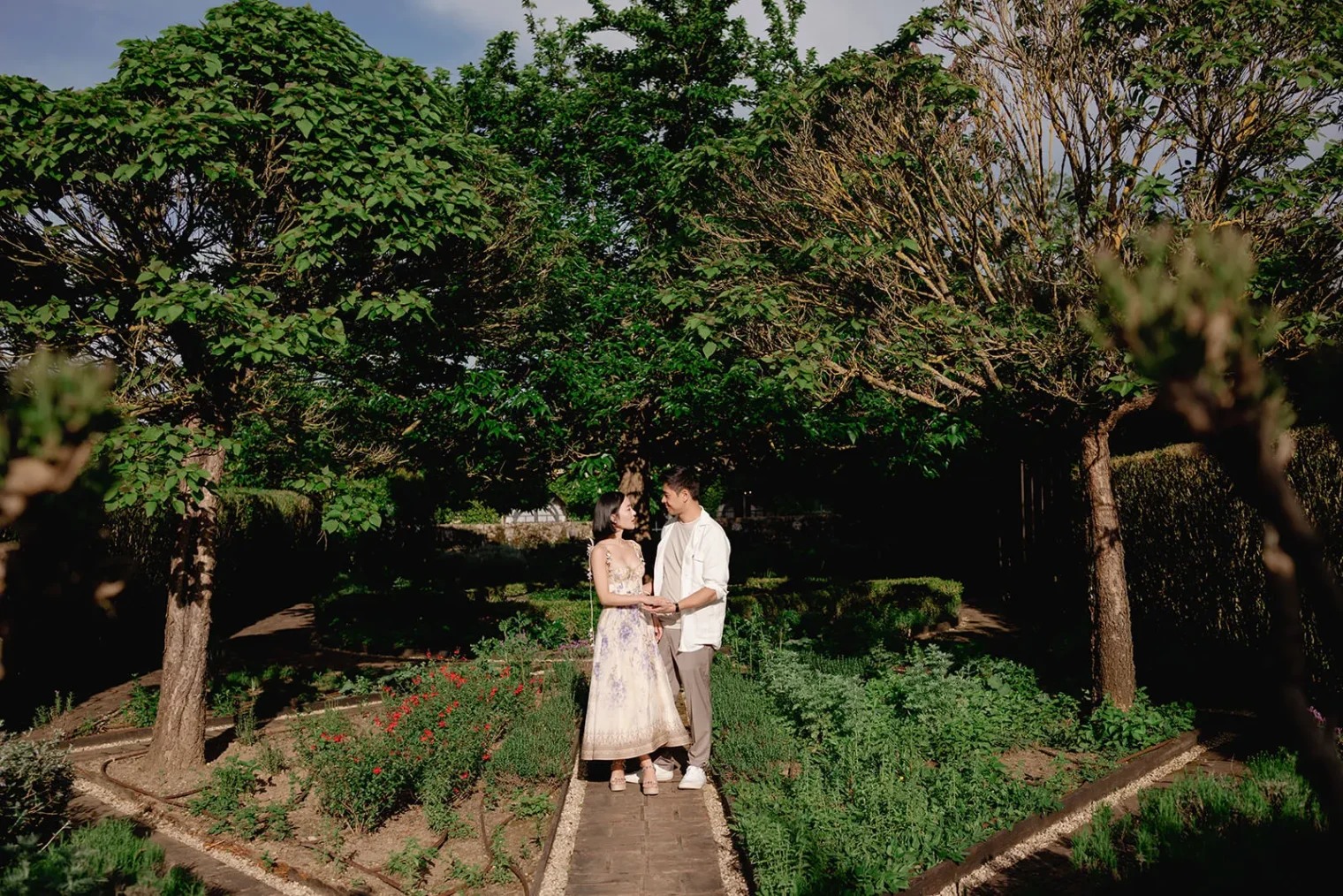 A couple stands holding hands on a garden path, surrounded by lush green plants and trees. Sunlight filters through the branches, casting dappled shadows. The woman wears a light floral dress; the man wears a white shirt and beige pants. Both look at each other, smiling. Tuscan Wedding Photographer