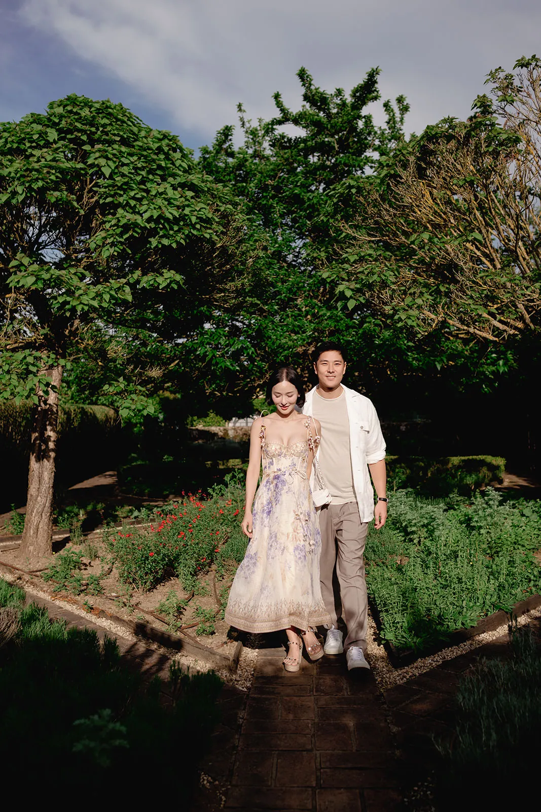 A woman in a light, floral dress and a man in a white shirt and gray pants stand side by side, smiling in a lush garden with tall green trees and sunlight casting soft shadows around them. Tuscan Wedding Photographer