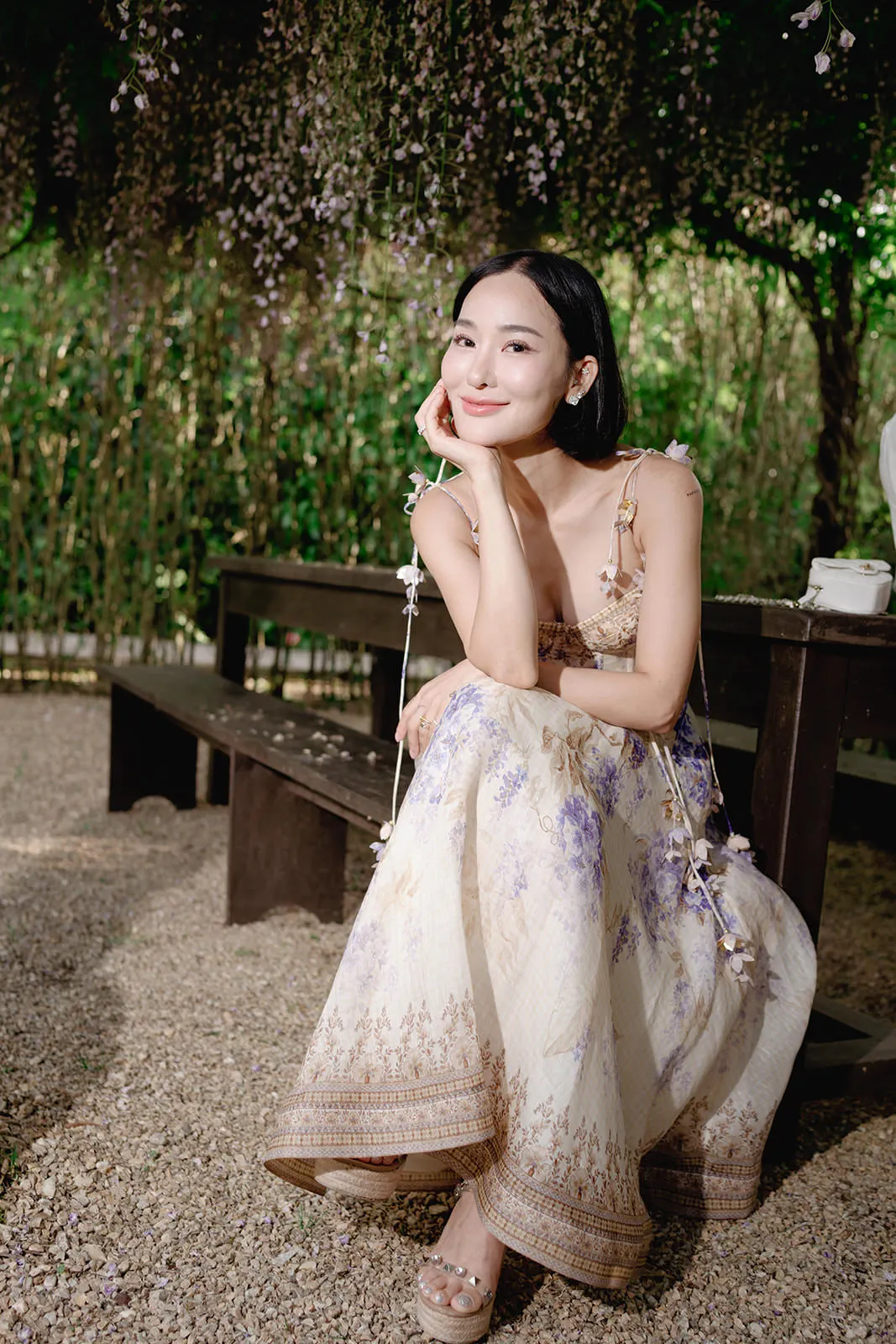 A woman with straight black hair sits on a wooden bench outdoors, surrounded by hanging flowers and green foliage. She wears a long, floral-patterned dress in cream and lavender tones, smiles gently, and rests her chin on one hand. Pebbles cover the ground. Tuscan Wedding Photographer