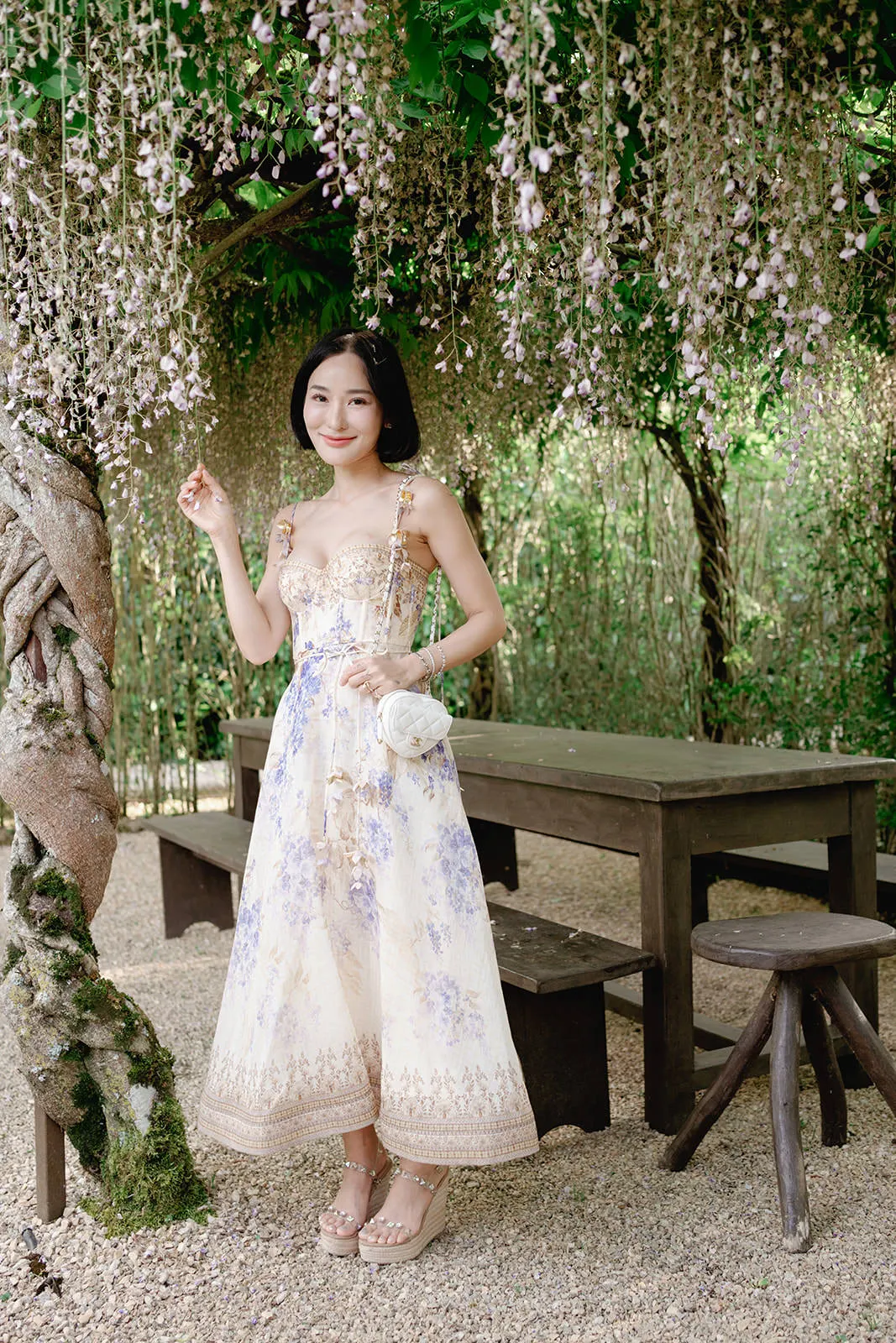 A woman in a light floral dress and sandals stands smiling under hanging wisteria flowers. She holds a white round purse and poses beside a rustic wooden table and benches, surrounded by lush greenery and a tree wrapped in vines on a gravel path. Tuscan Wedding Photographer