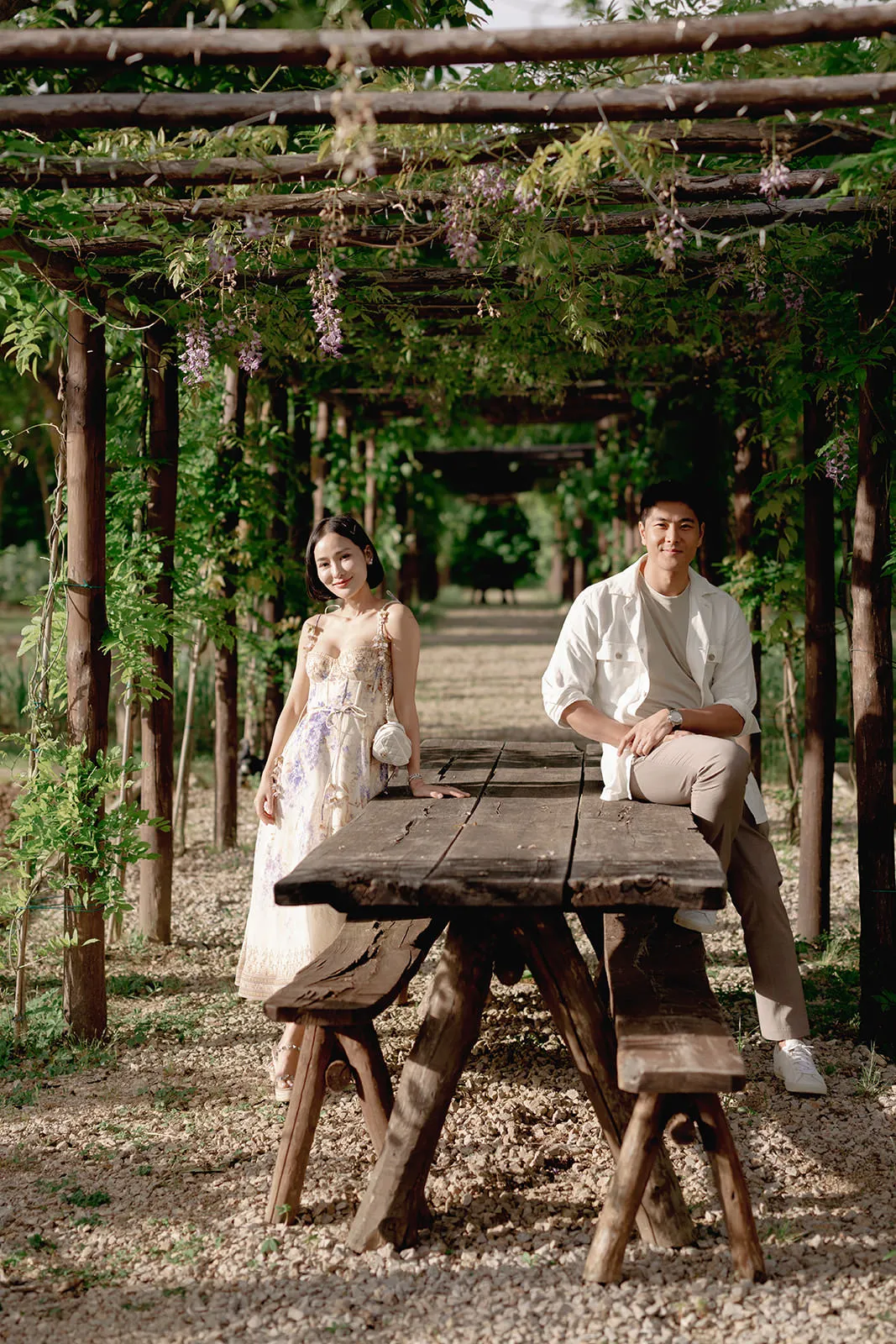 A woman in a floral dress stands at one end of a rustic wooden table under a pergola covered with green vines, while a man in a white shirt and beige pants sits on the table at the other end. Sunlight filters through, casting natural light and shadows. Tuscan Wedding Photographer