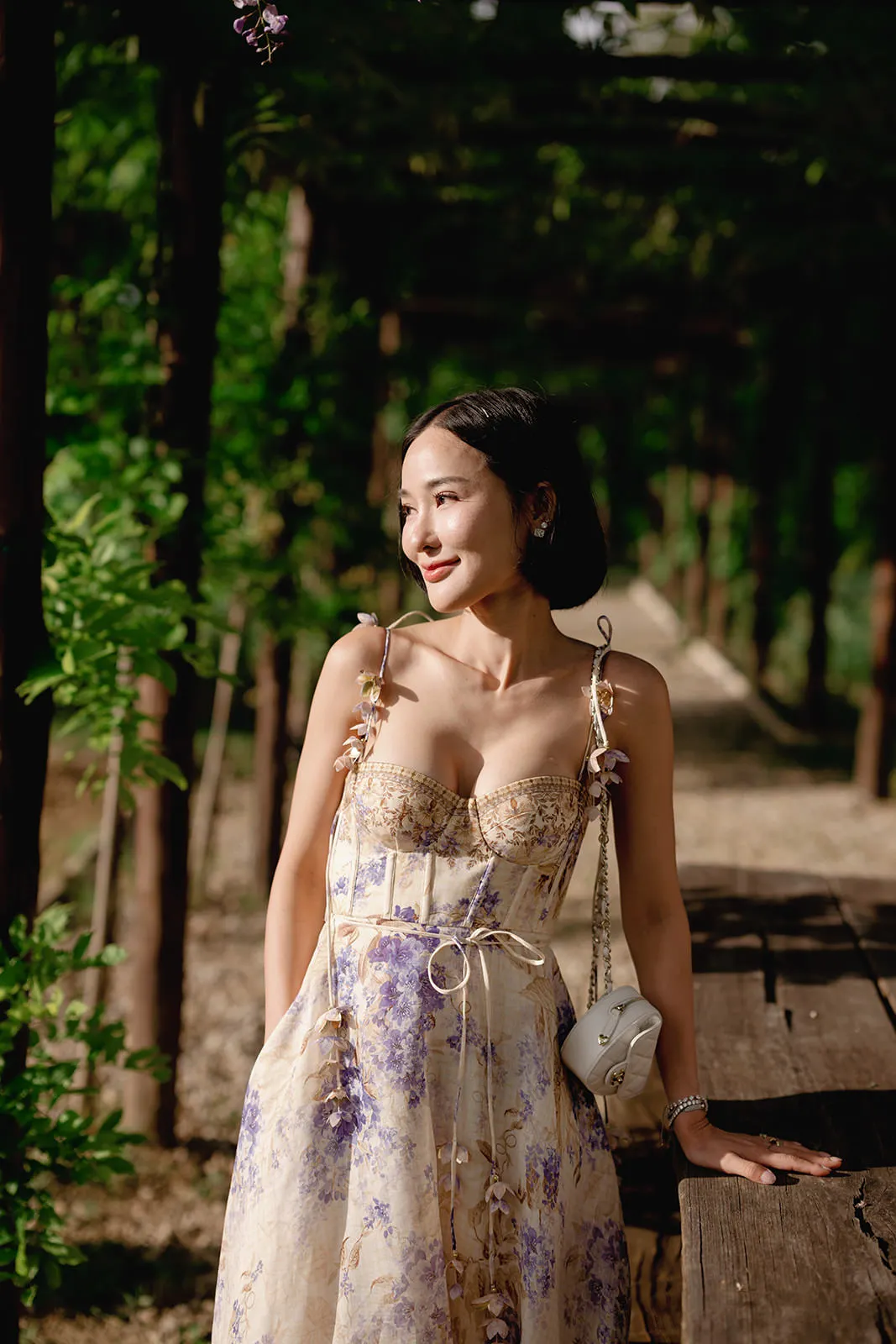 A woman with short dark hair stands outdoors under a pergola, wearing a light yellow floral dress with purple patterns. She holds a small white purse with a chain strap and smiles slightly, looking to her left. Sunlight shines on her and green foliage surrounds her. Tuscan Wedding Photographer