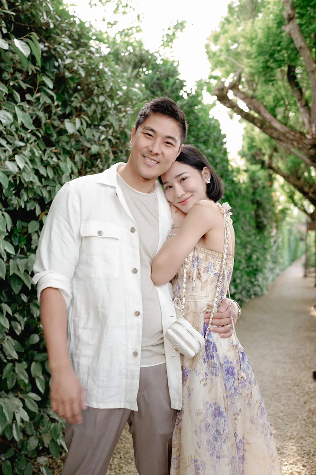 A smiling couple poses on a leafy outdoor path. The man wears a white shirt over a beige t-shirt and gray pants. The woman, in a floral dress with a white purse across her body, hugs him with her head on his chest. Sunlight filters through the trees. Tuscan Wedding Photographer