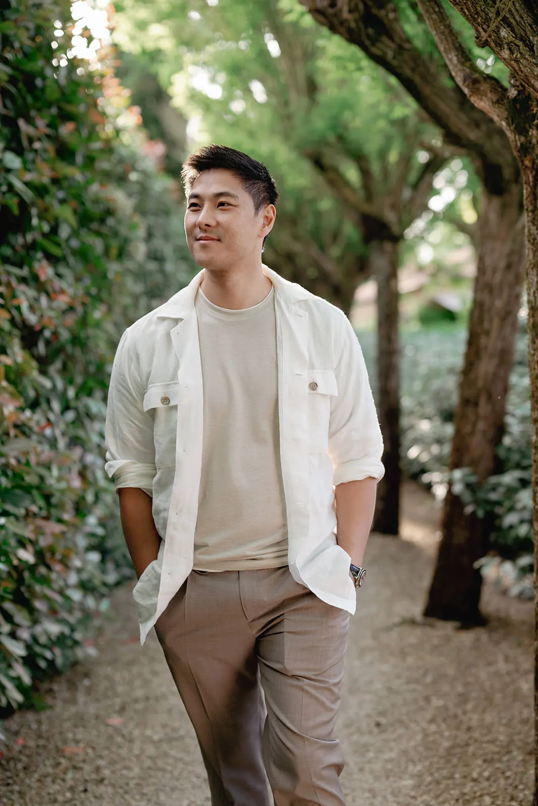 A man with short black hair, wearing a white button-up shirt over a beige t-shirt and light brown pants, walks outside on a gravel path lined with green bushes and trees, with hands in his pockets, smiling and looking to the side. Tuscan Wedding Photographer