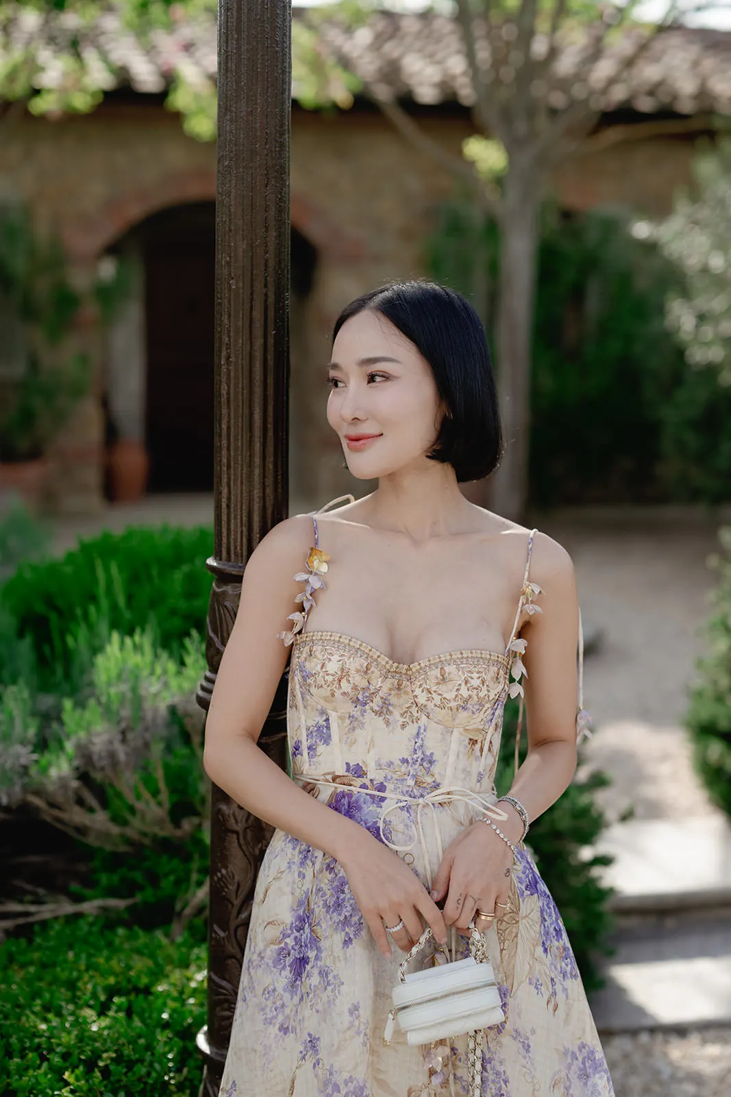A woman with a short black bob stands outdoors by a dark post, wearing a floral-patterned, sleeveless dress with thin straps and flower details. She holds a small white purse, looking to the side and smiling slightly, with greenery and a rustic building behind her. Tuscan Wedding Photographer