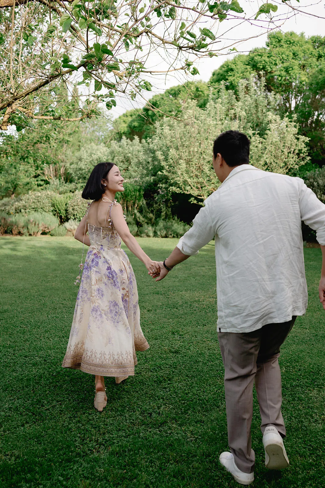 A woman in a floral dress smiles as she leads a man by the hand across a grassy lawn. The man wears a light shirt and trousers. They walk towards lush green trees and plants on a bright, sunny day, suggesting a joyful and romantic moment outdoors. Tuscan Wedding Photographer