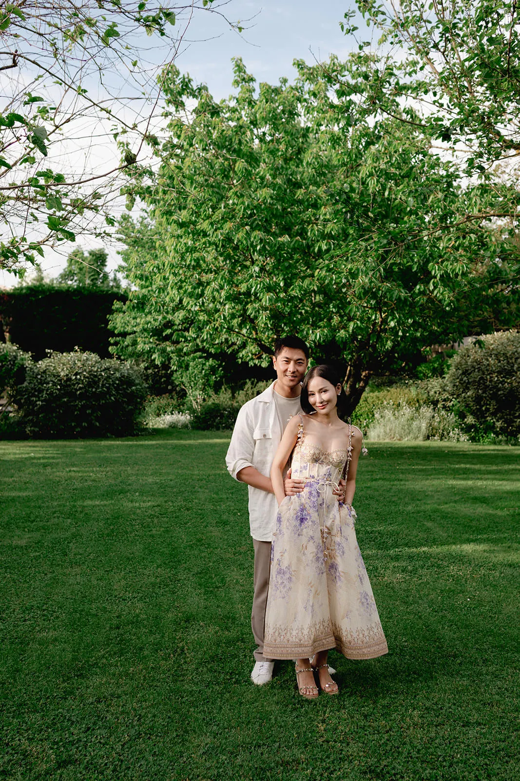 A young couple stands on a lush green lawn, surrounded by trees and shrubs. The man, wearing a white shirt and beige pants, embraces the woman from behind. She wears a long, sleeveless, cream dress with purple floral details. Both smile gently at the camera. Tuscan Wedding Photographer