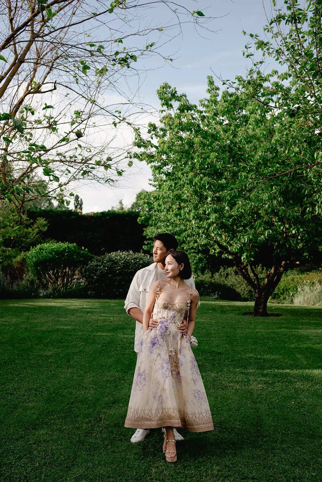 A man in a white shirt stands behind a woman in a long, floral, sleeveless dress. They are on a green lawn with trees and shrubs in the background. The woman has her hands in her pockets, and both look off to the side under a partly cloudy sky. Tuscan Wedding Photographer
