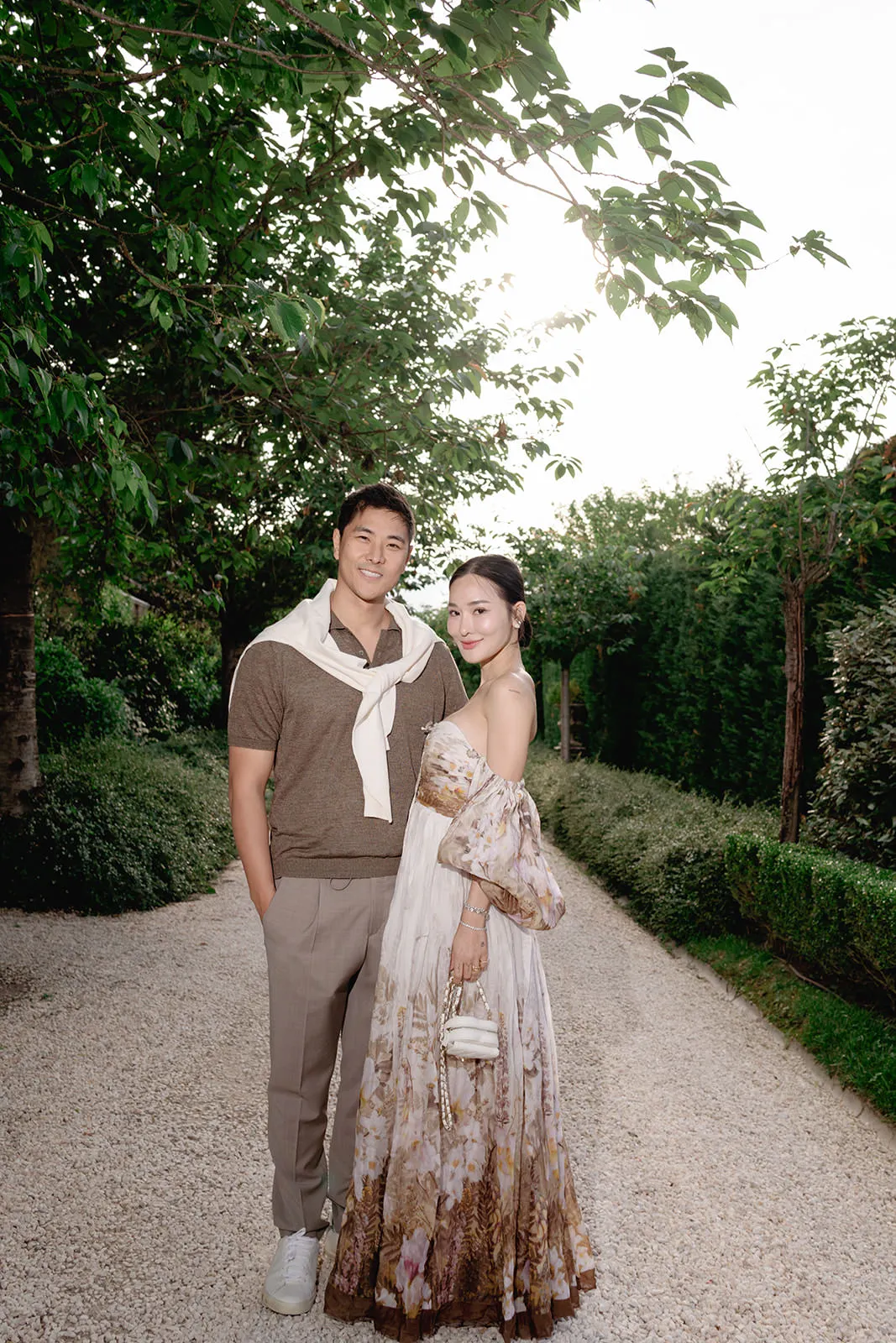 A smiling couple stands on a gravel path lined with greenery. The man wears a brown polo, beige pants, white sneakers, and a cream sweater draped over his shoulders. The woman wears an off-the-shoulder floral maxi dress and holds a small white bag. Trees frame the scene. Tuscan Wedding Photographer