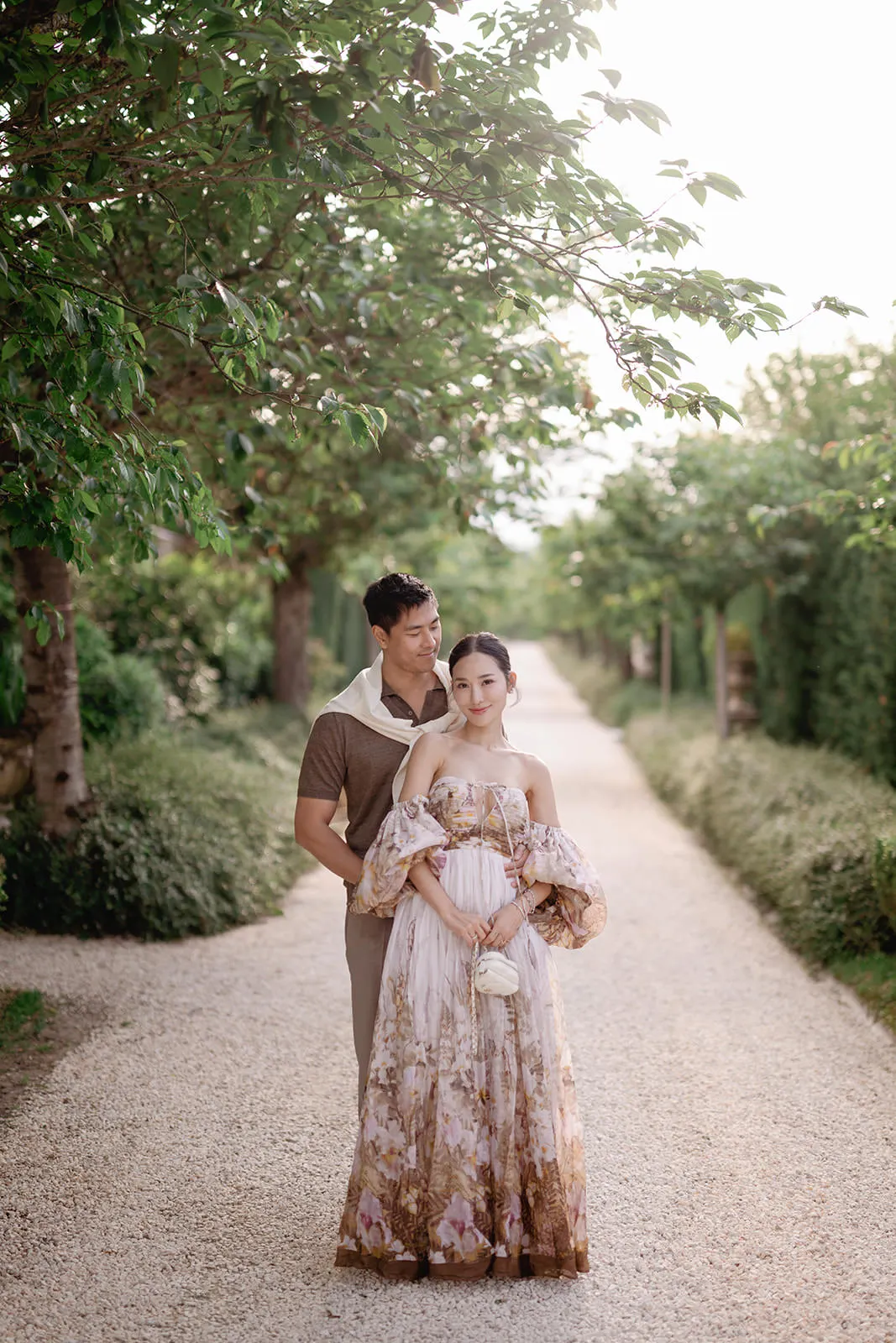 A couple poses on a tree-lined gravel path. The man, in a brown shirt and cream sweater, stands behind the woman, who wears a flowing, off-shoulder floral dress and holds a small bouquet. Sunlight filters gently through the trees, creating a soft, romantic scene. Tuscan Wedding Photographer