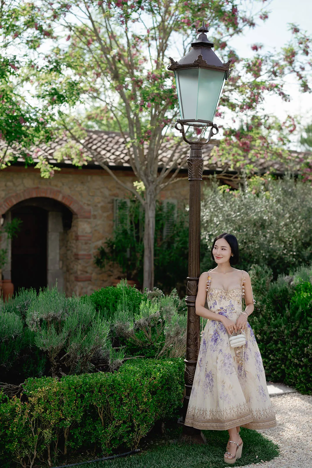 A woman in a light floral dress stands by a tall vintage lamp post in a lush garden. She holds a small white purse and wears sandals. Behind her, trimmed bushes, lavender, and trees surround a rustic stone house with a tiled roof and arched doorway. Tuscan Wedding Photographer