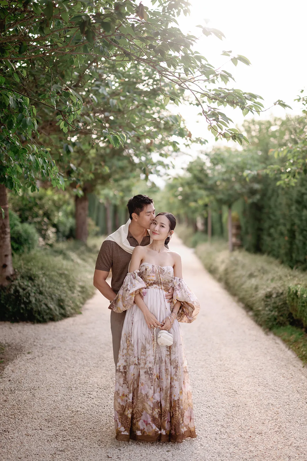 A couple stands on a sunlit gravel path lined with green trees. The man hugs the woman from behind, gently kissing her head—a perfect moment often captured in our Wedding Photography Packages for couples seeking timeless memories. Tuscan Wedding Photographer