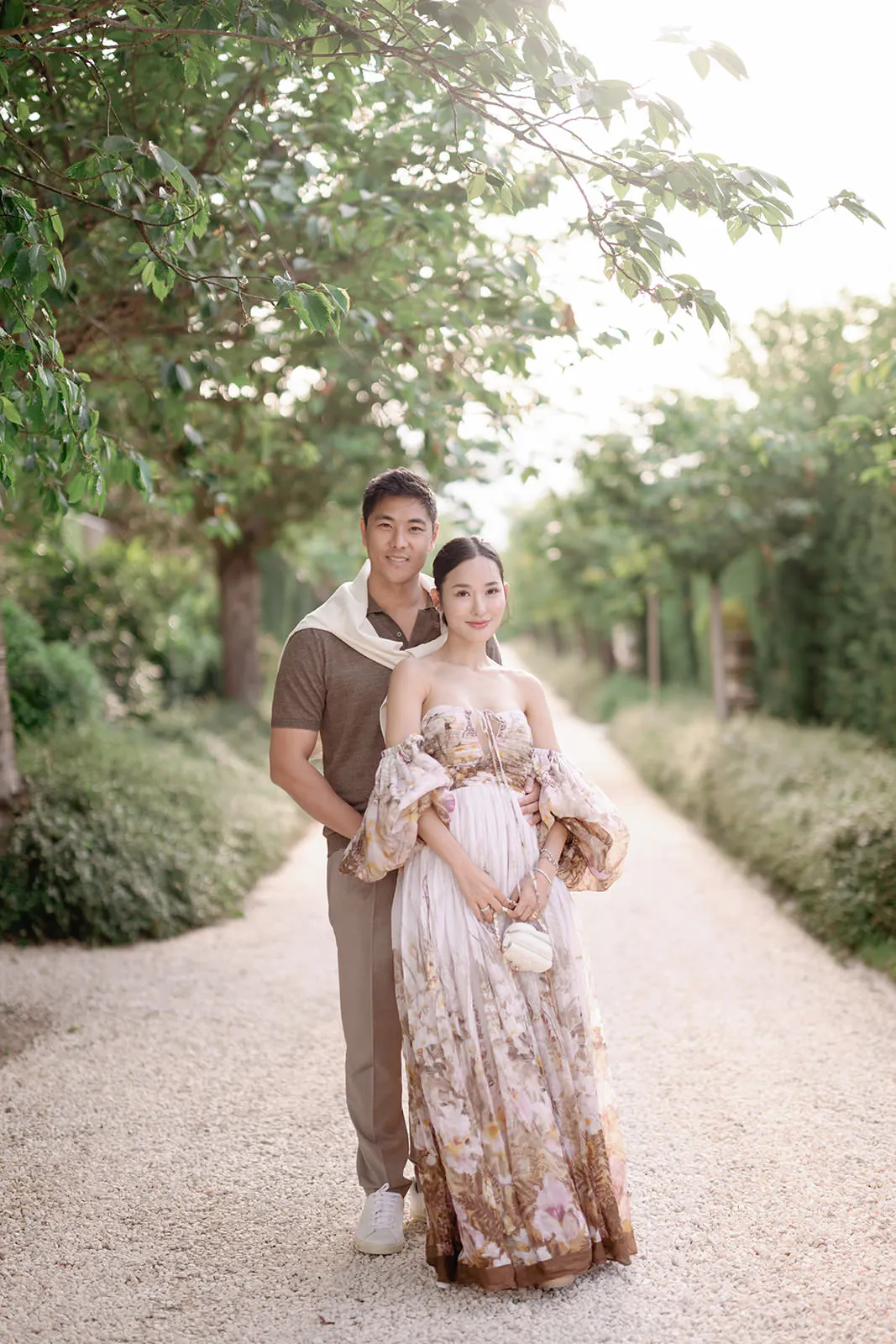 A man and a woman stand on a sunlit gravel path lined with green trees and bushes. The man, in a brown shirt and beige pants, stands behind the woman, who wears a floral off-shoulder dress and holds a small purse. Both smile softly at the camera. Tuscan Wedding Photographer