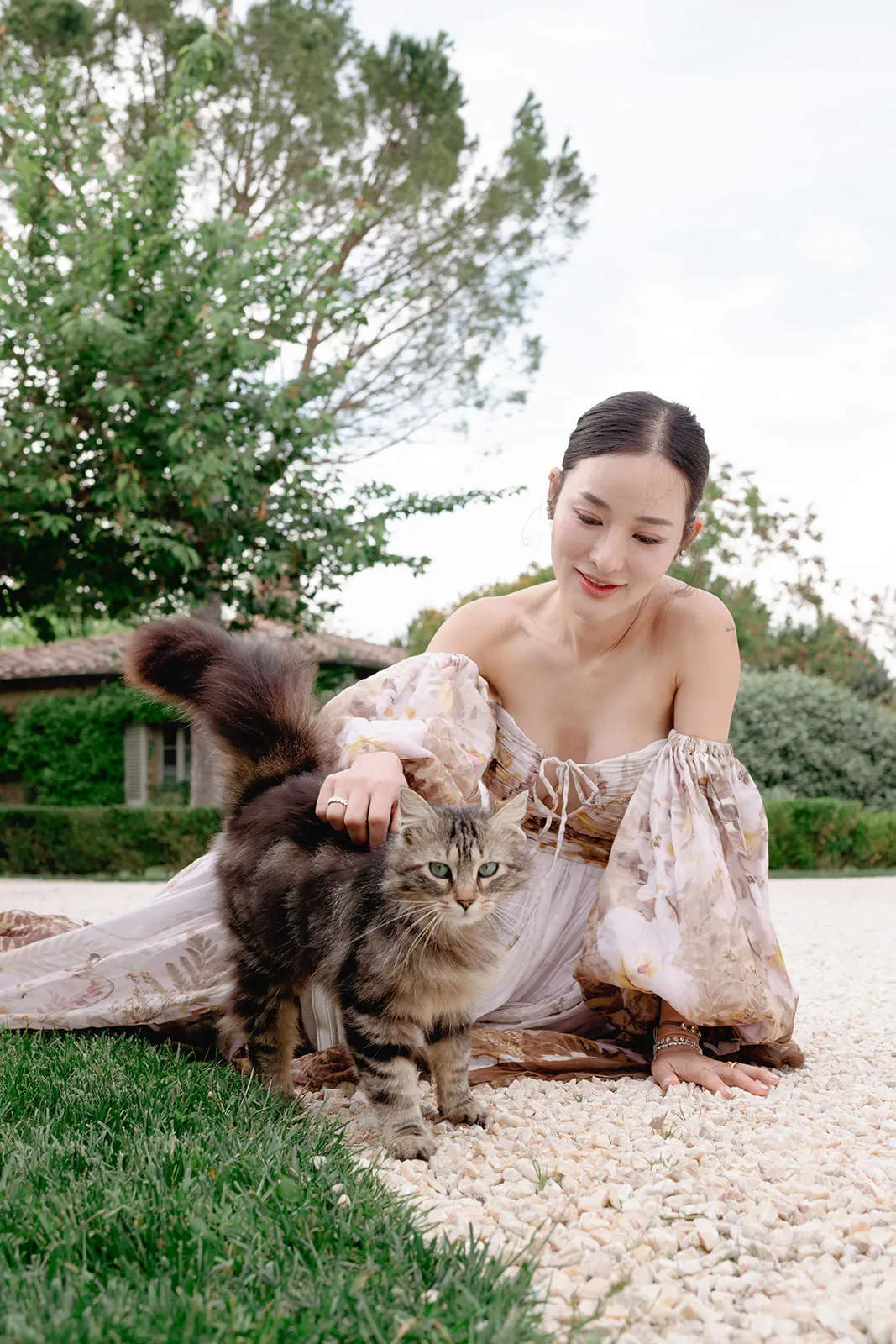 A woman in a flowy, floral off-shoulder dress kneels on a stone path, gently petting a fluffy, long-haired tabby cat. She smiles warmly at the cat. Green grass and leafy trees are in the background under a partly cloudy sky. Tuscan Wedding Photographer