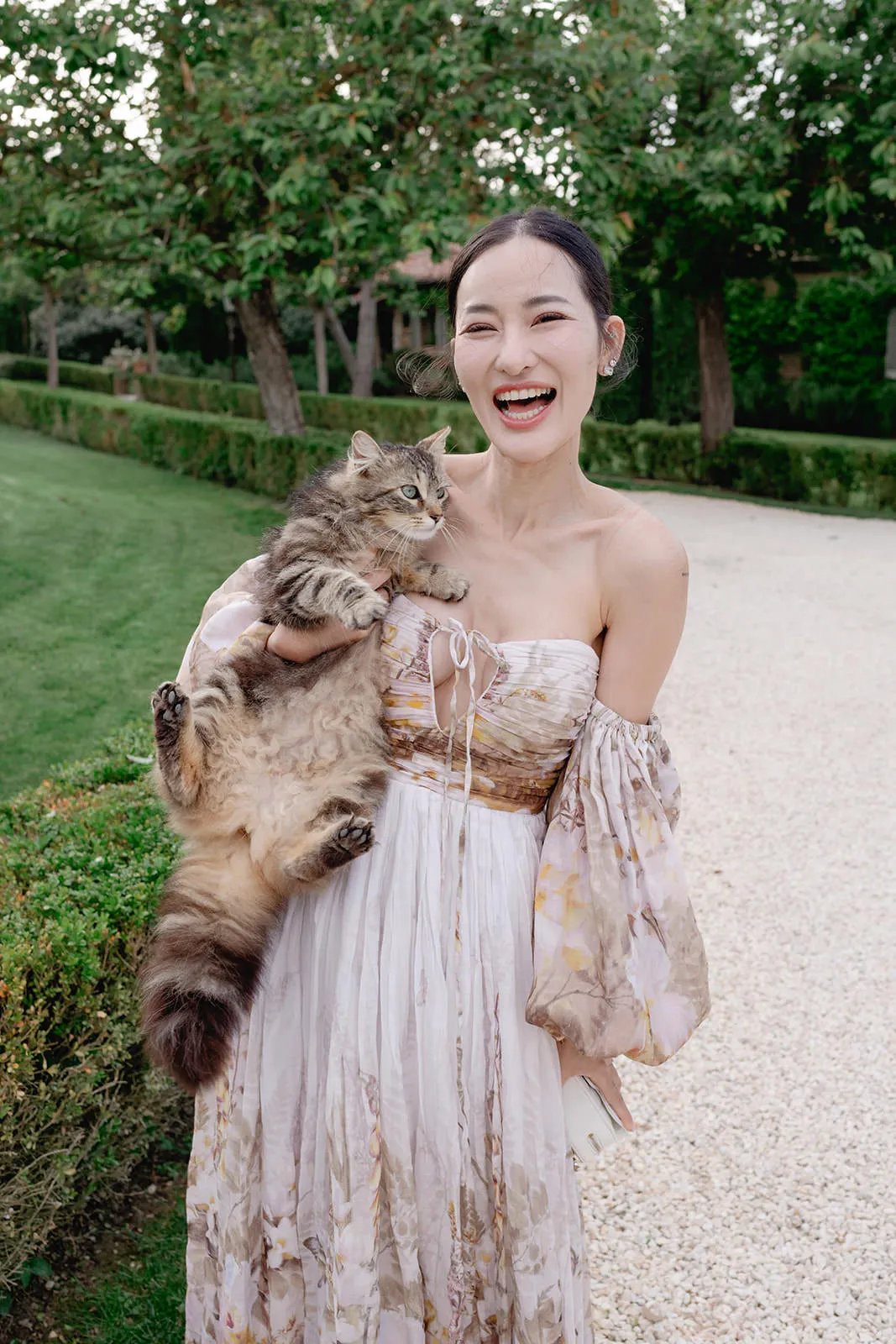 A smiling woman in a floral off-the-shoulder dress stands outside on a gravel path, holding a large fluffy tabby cat. Green hedges, grass, and trees are visible in the background, creating a serene garden setting. Tuscan Wedding Photographer