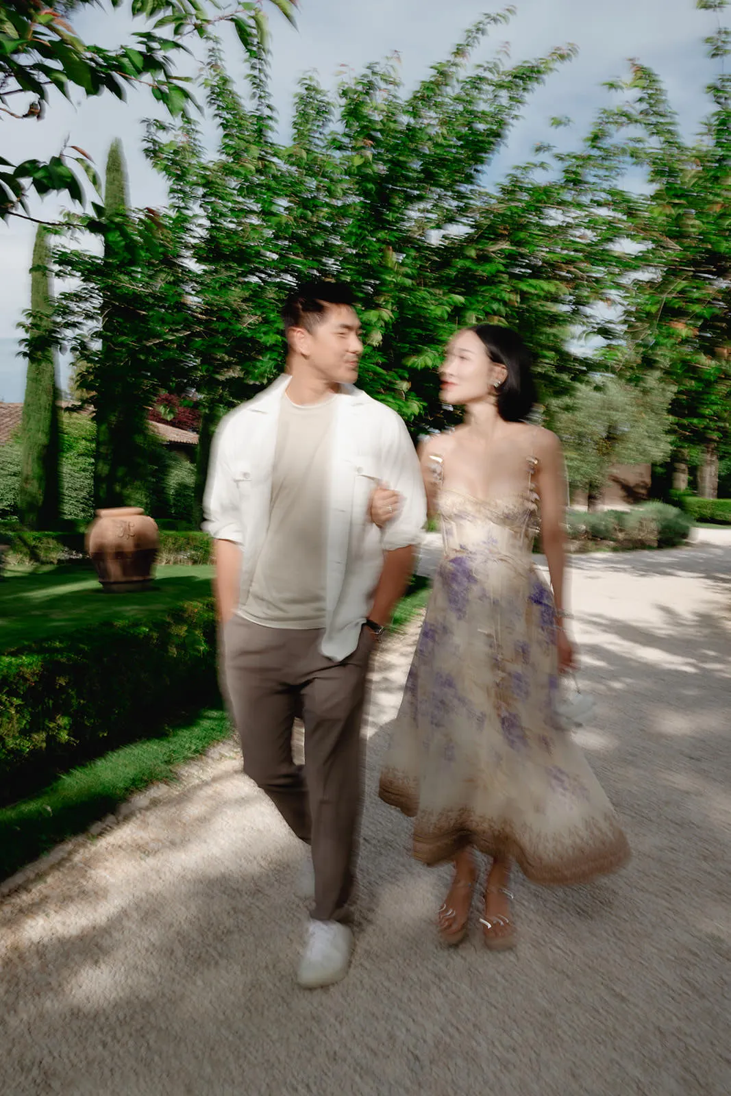 A man and woman walk arm-in-arm on a sunlit garden path, smiling at each other. The man wears a white shirt and khaki pants; the woman wears a light floral dress. Lush greenery, trees, and trimmed bushes surround them. The image is slightly blurred. Tuscan Wedding Photographer