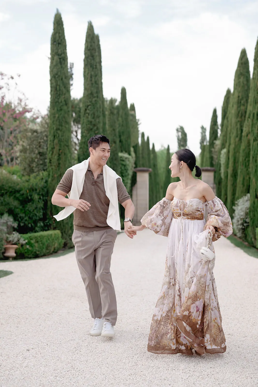 A smiling couple walks hand in hand down a gravel path lined with tall cypress trees. The man wears a brown shirt, light trousers, white vest, and sneakers. The woman wears a flowing off-shoulder floral dress and carries a small white purse. Both look joyful and relaxed. Tuscan Wedding Photographer