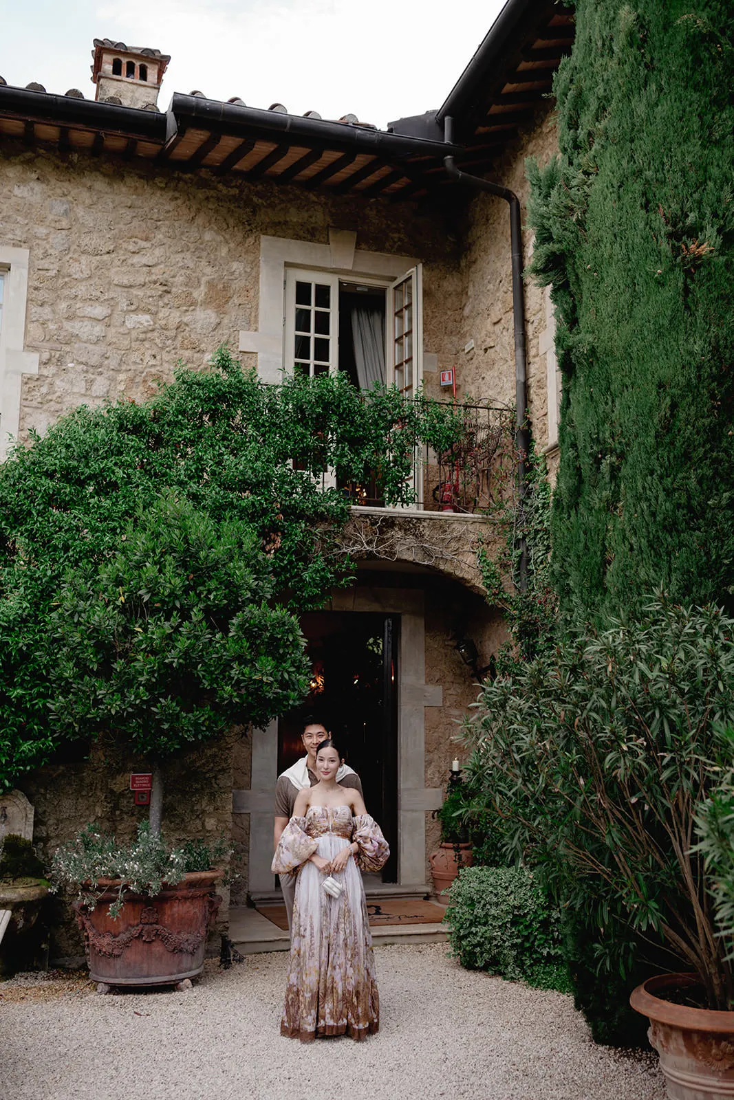 A couple stands in front of a rustic stone building covered in green vines. The woman wears a long, patterned dress and the man stands behind her, holding her waist. Tall greenery and potted plants surround them, creating a romantic, serene outdoor setting. Tuscan Wedding Photographer