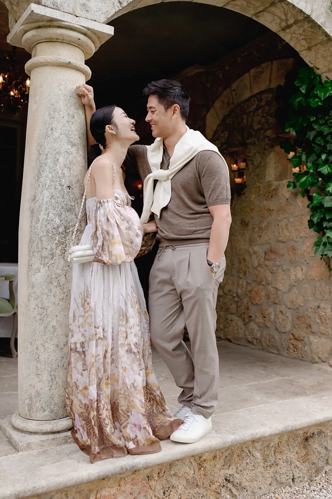 A smiling couple stands close under a stone archway. The woman wears a flowing, off-shoulder floral dress and carries a white bag. The man wears a brown shirt, taupe pants, white sneakers, and a white sweater draped over his shoulders. Both look happy and affectionate. Tuscan Wedding Photographer