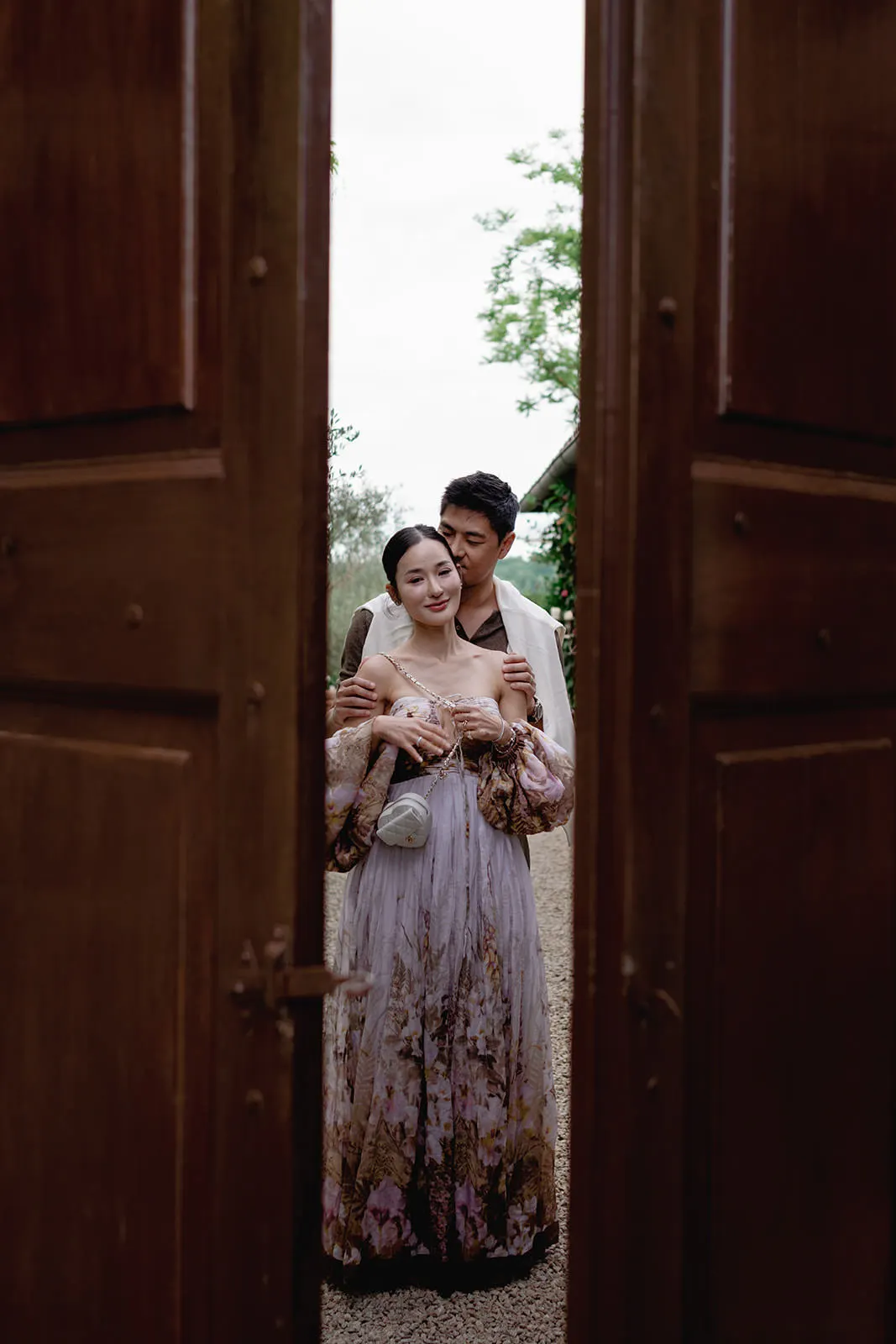 A couple stands closely together, framed between two large wooden doors. The woman wears an off-shoulder, flowy, floral dress and smiles gently while the man stands behind, holding her with his arms around her waist. Greenery is visible in the background. Tuscan Wedding Photographer