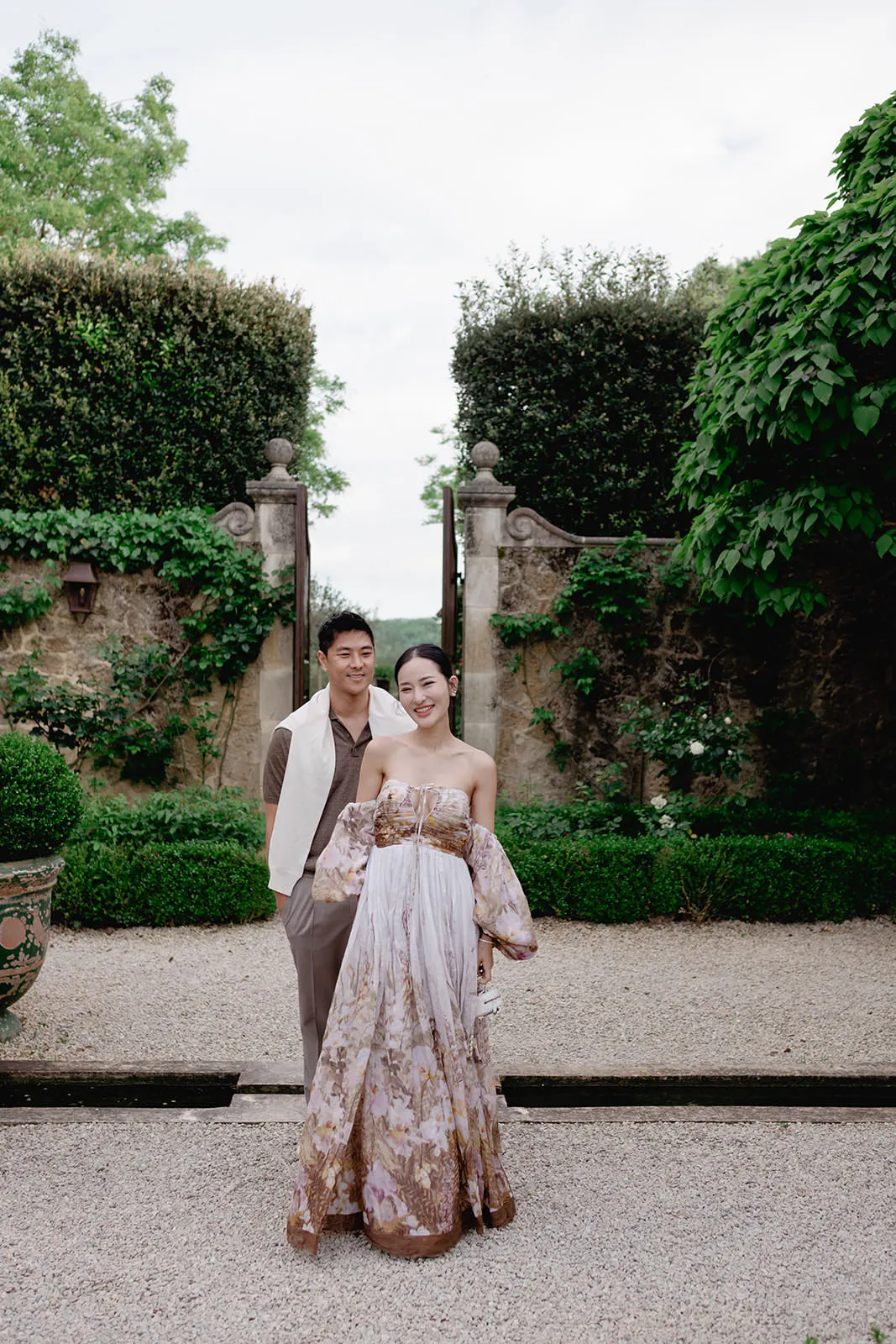 A couple stands outdoors by a stone gate surrounded by greenery. The woman wears a flowing, off-the-shoulder floral dress; the man stands slightly behind her in a tan outfit with a white jacket draped over his shoulder. They both smile at the camera. Tuscan Wedding Photographer