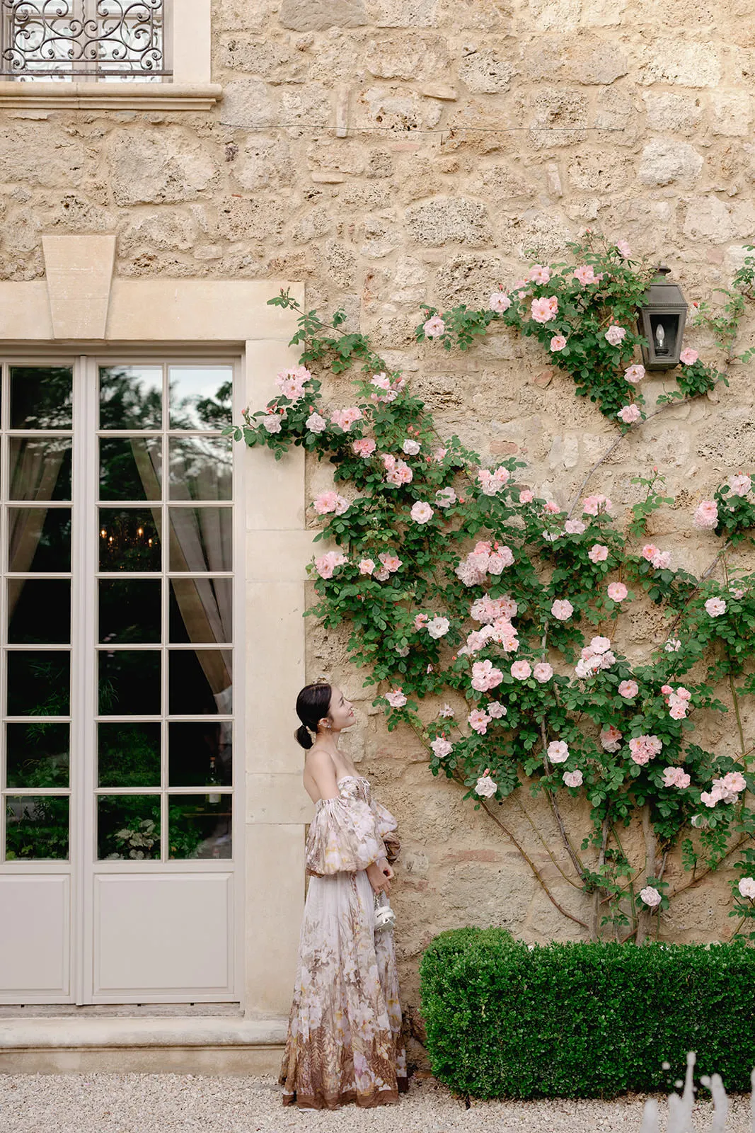A woman in a flowing, floral off-shoulder dress stands beside a beige stone wall covered with blooming pink roses. She looks up at the flowers. The wall has a tall window with sheer curtains and a black lantern above the rose bush. Tuscan Wedding Photographer