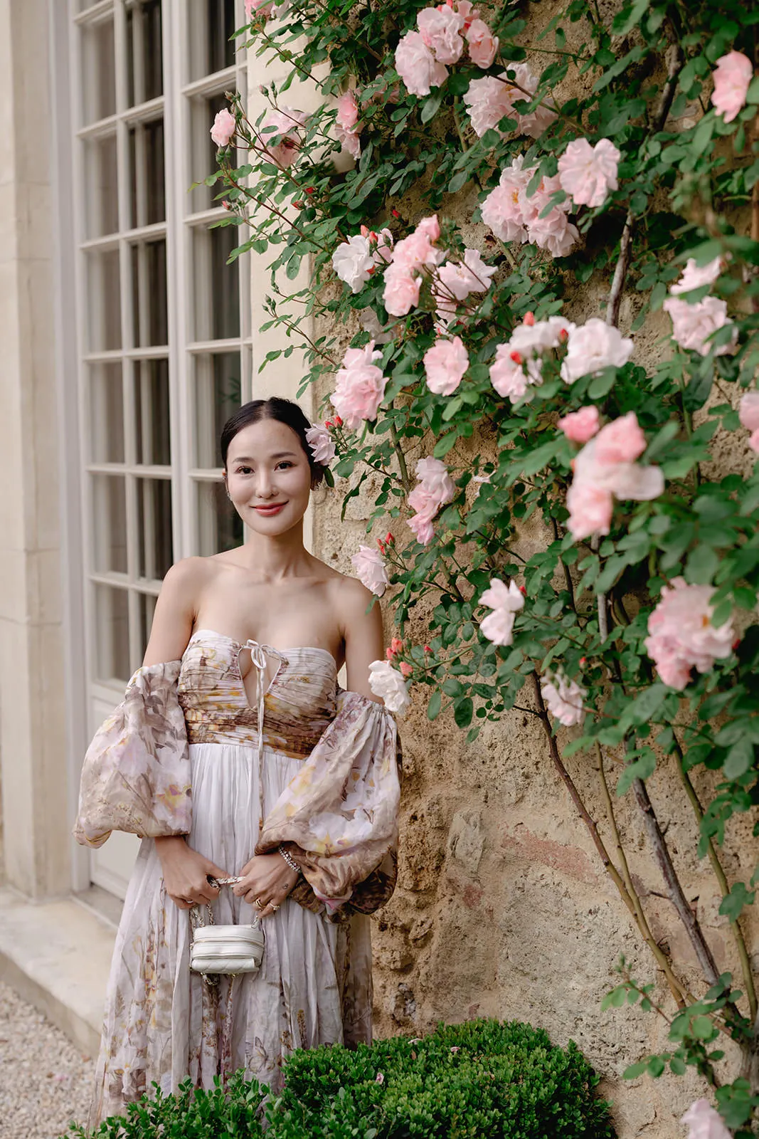A woman in an off-the-shoulder, floral-patterned dress stands beside a stone wall covered in blooming pink roses. She holds a small silver purse, has dark hair pulled back, and smiles gently. A large window and green shrubbery are also visible. Tuscan Wedding Photographer