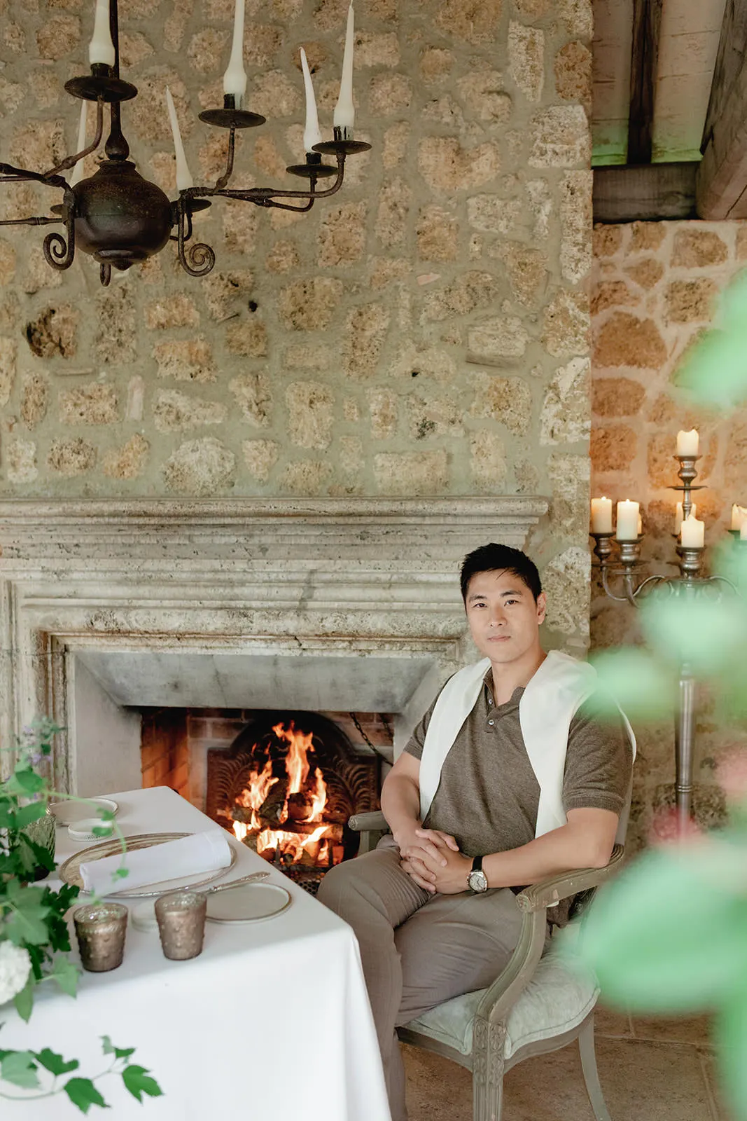 A man sits at a dining table covered with a white cloth, near a stone fireplace with a fire burning. He wears a brown shirt and a light sweater draped over his shoulders. The rustic room has stone walls, candles, a chandelier, and green plants decorate the table. Tuscan Wedding Photographer