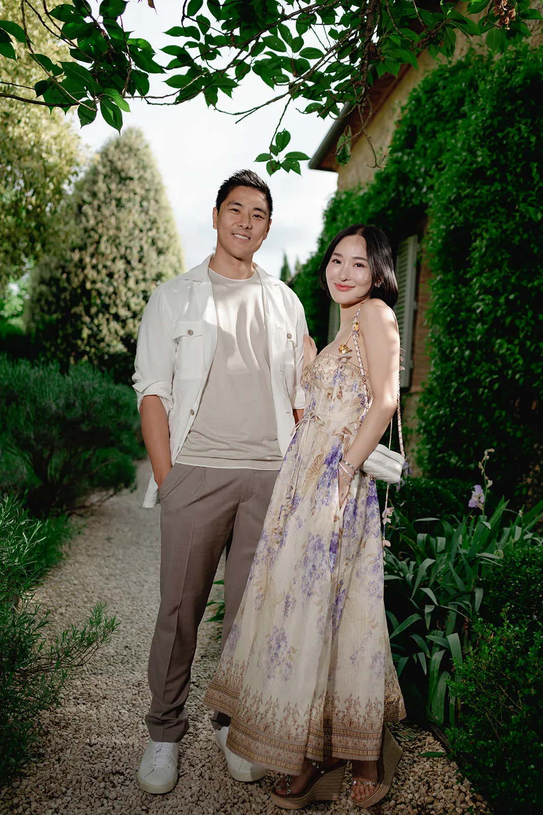 A smiling man in a white jacket and beige pants stands beside a woman in a long, sleeveless floral dress. They are outdoors on a gravel path, surrounded by green plants and bushes, with a house covered in ivy in the background. Tuscan Wedding Photographer